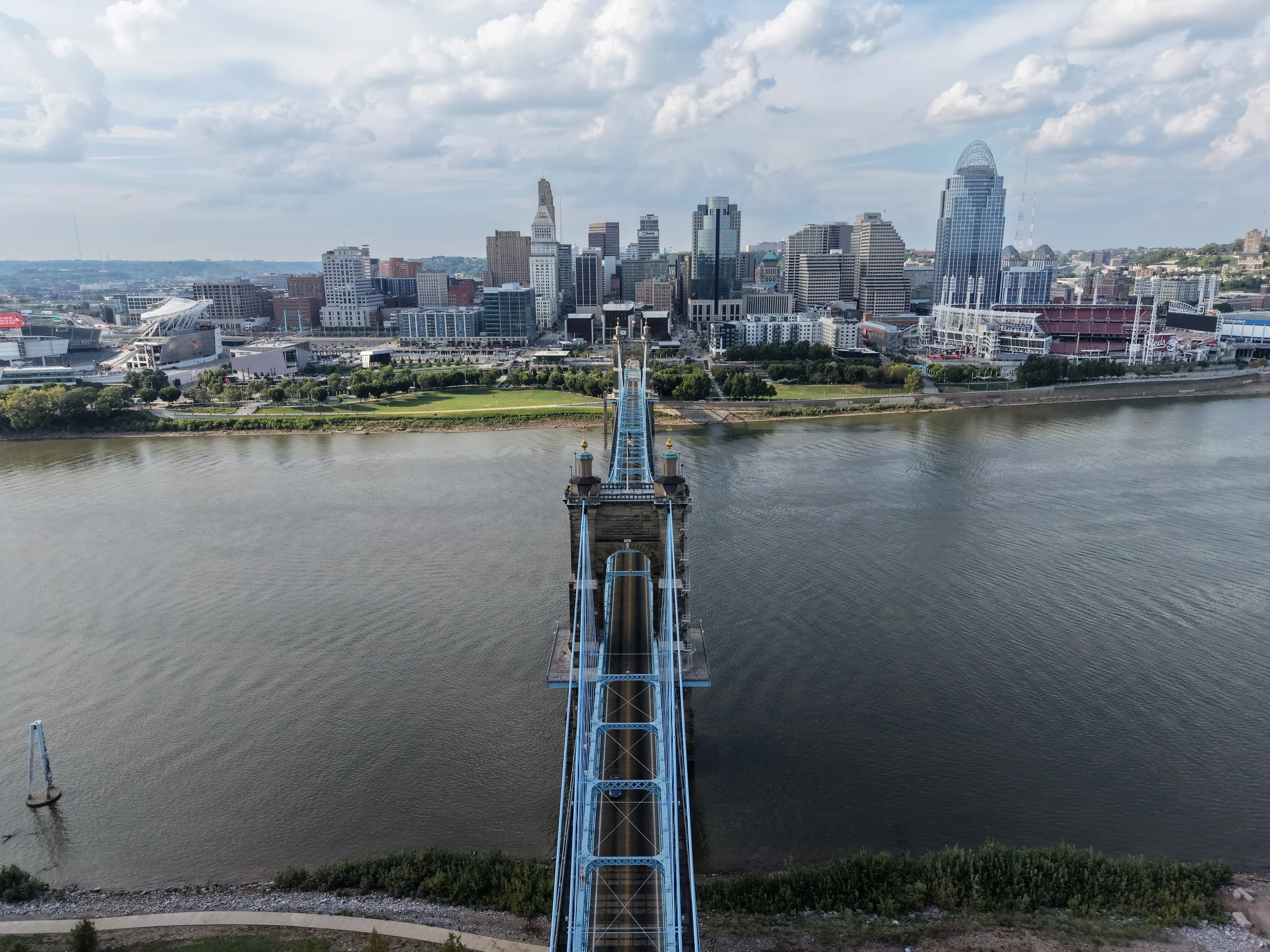 Aerial view of a city skyline with tall buildings and a river in the foreground. A bridge extending over the river connects to the city, with a stadium on the right side of the image. The sky is partly cloudy.