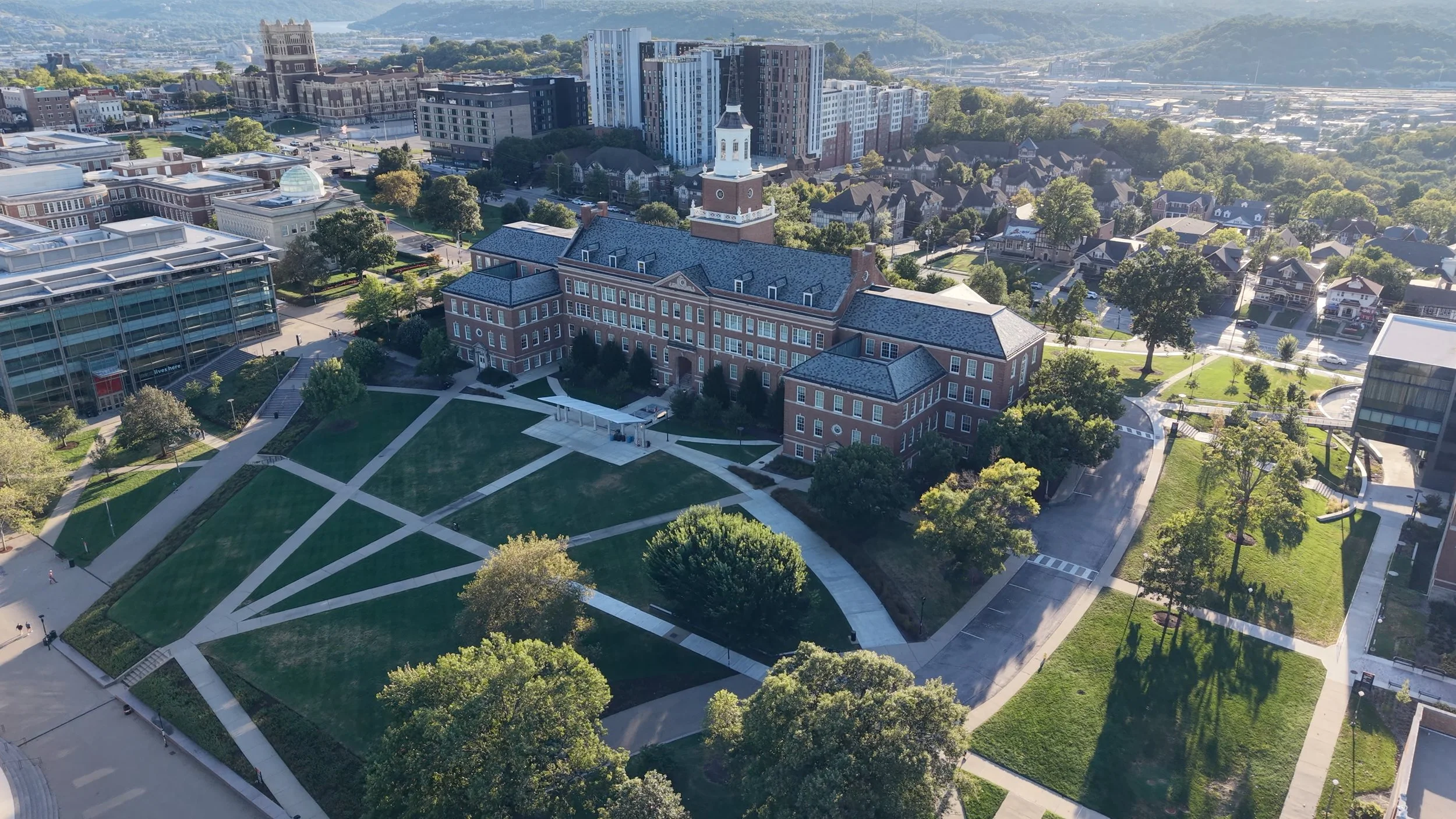 Bird's eye view of a college campus with a prominent red brick building featuring a clock tower, surrounded by green lawn areas, trees, walkways, and modern high-rise buildings in the background.