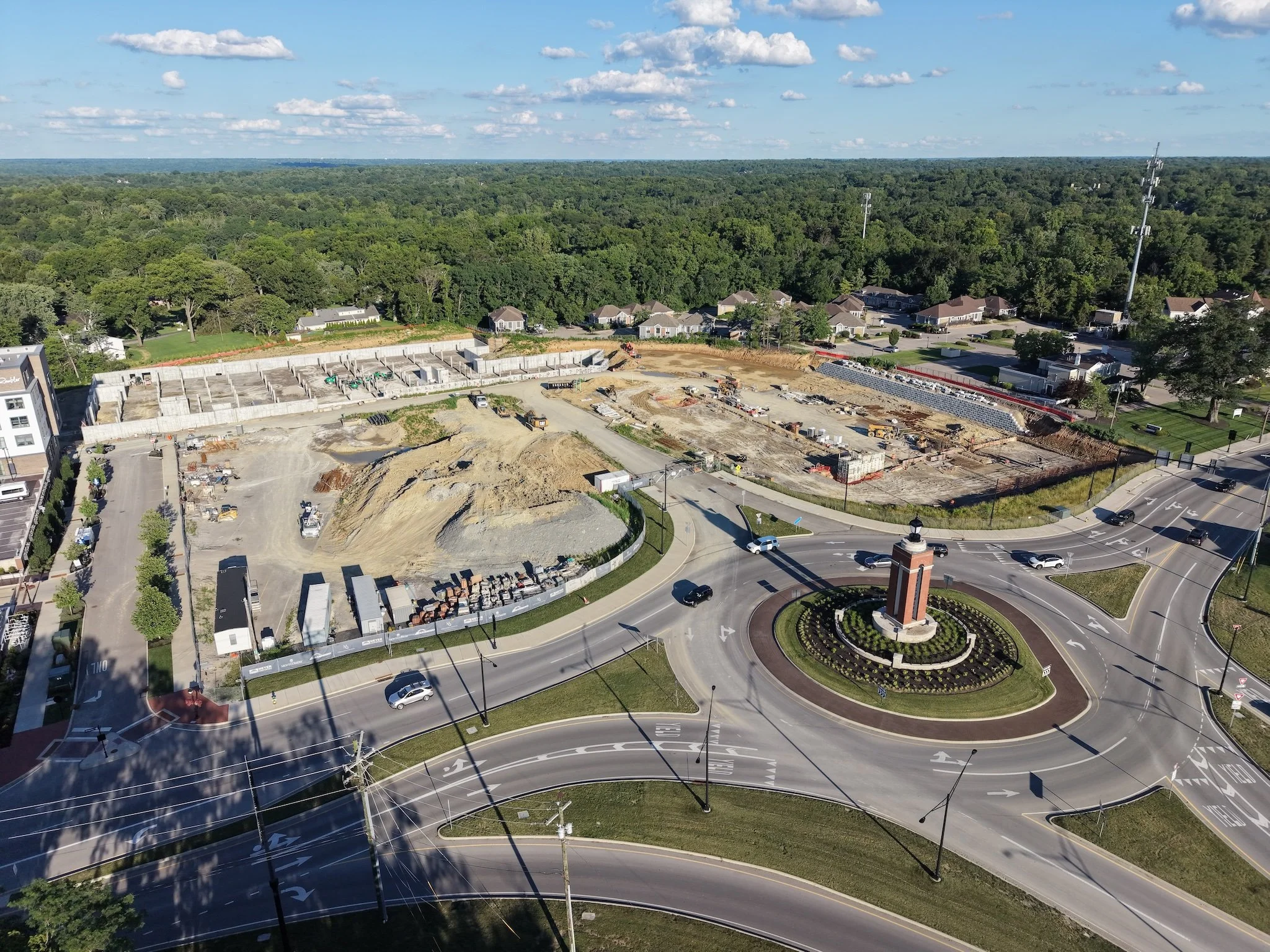 A drone aerial view of a construction site beside a busy road with multiple lanes, cars, and a roundabout; surrounded by trees and residential houses with a forest in the background under a partly cloudy sky.