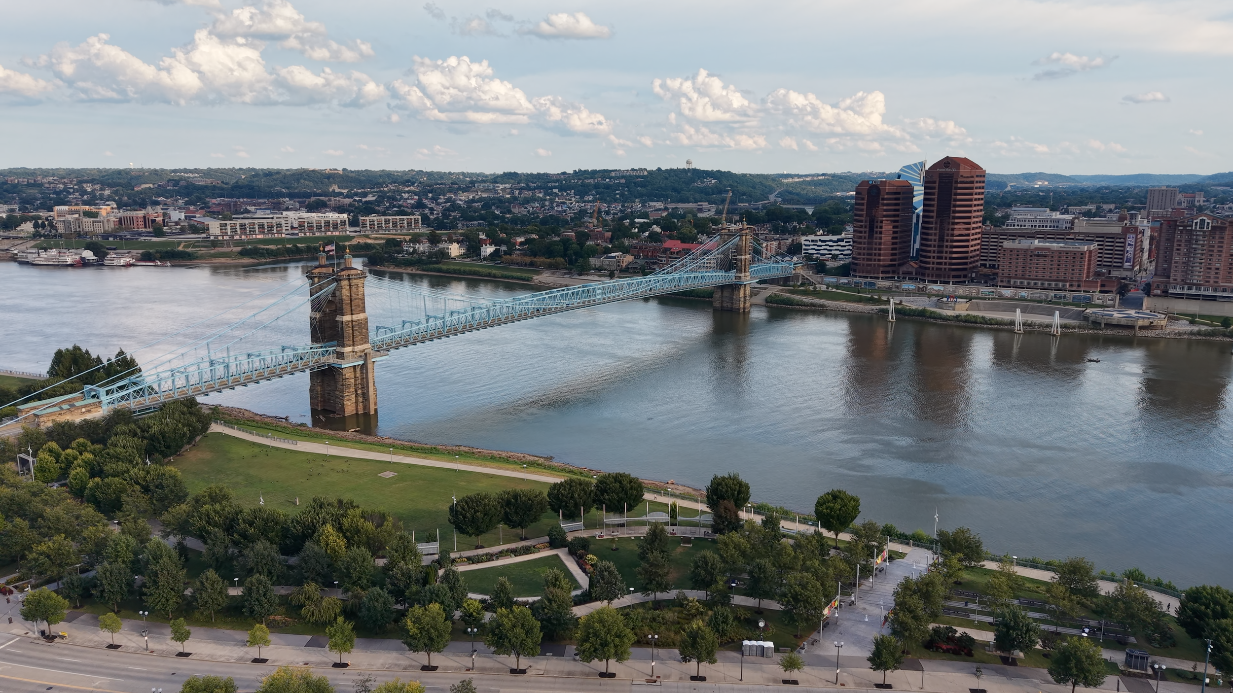 Aerial view of a city with a river, a suspension bridge, high-rise buildings, and a park with green trees.