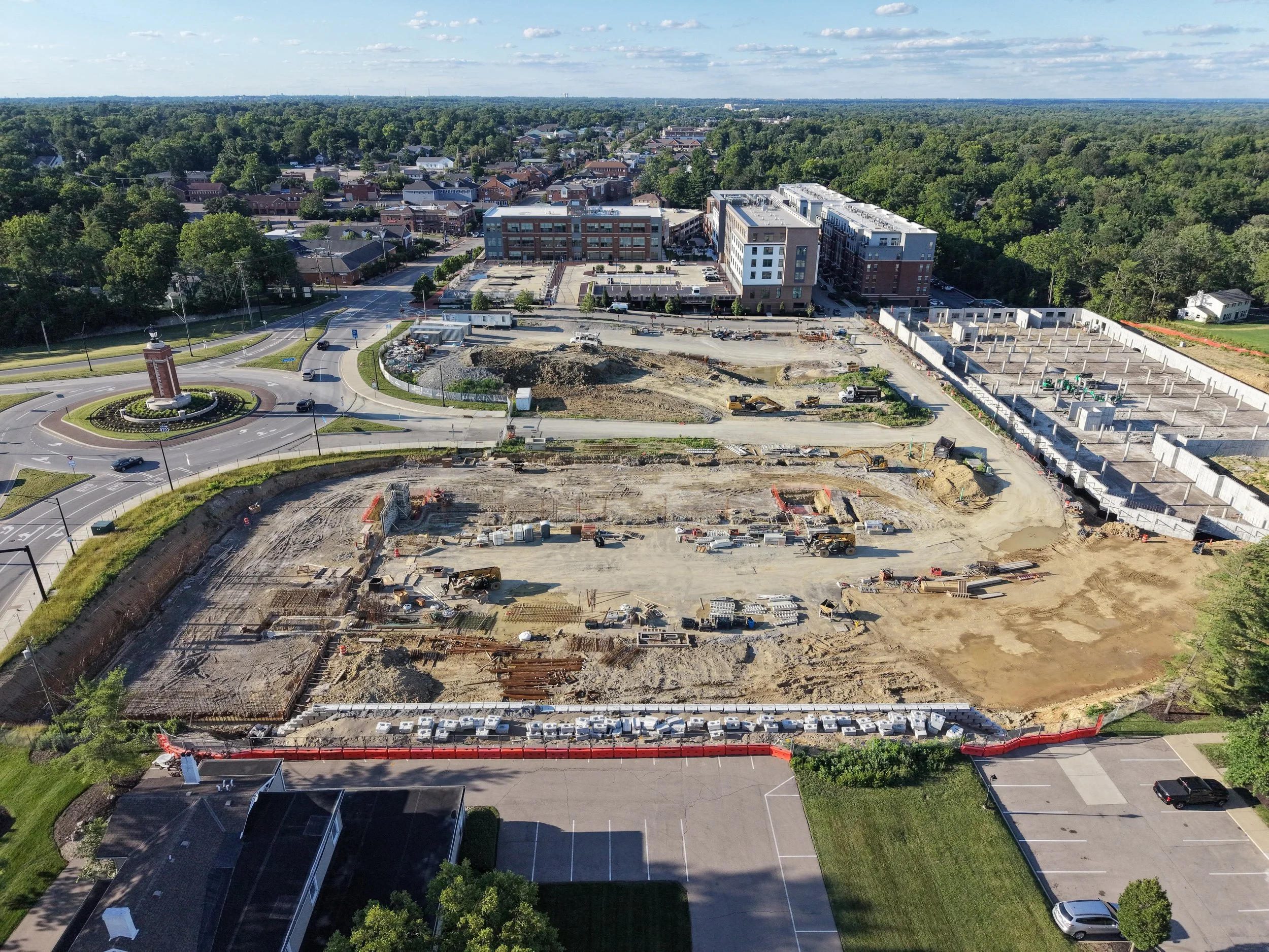 Aerial view of a construction site with cleared land, construction machinery, and partially built structures, surrounded by trees and residential neighborhood.