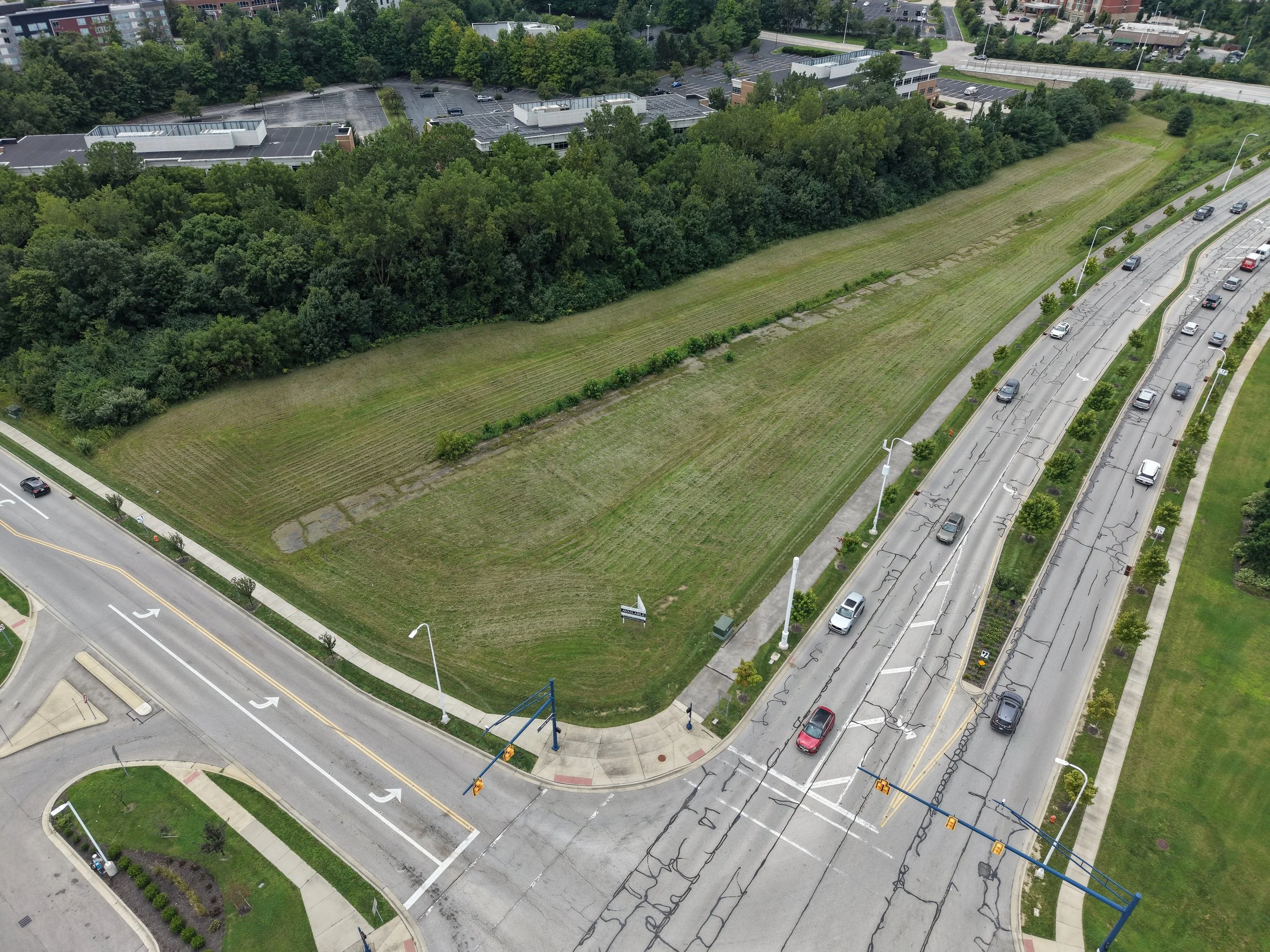 Aerial view of a parking lot with empty spaces, surrounded by trees and a grassy field, near a road with cars and streetlights.