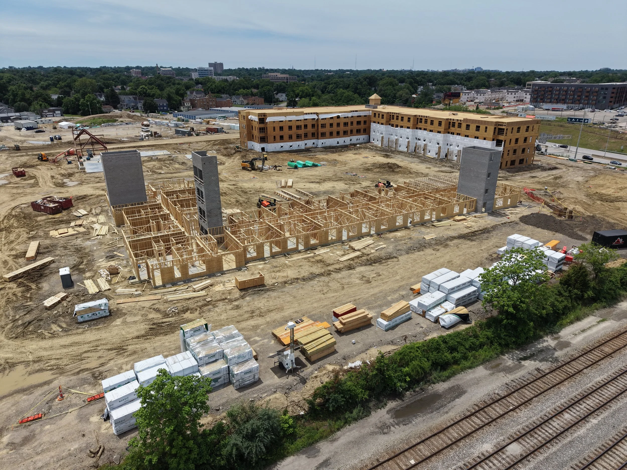Construction site with partially built multi-story building and wooden framework. Construction materials and equipment are scattered around, with a nearby railroad track and residential area in the background.