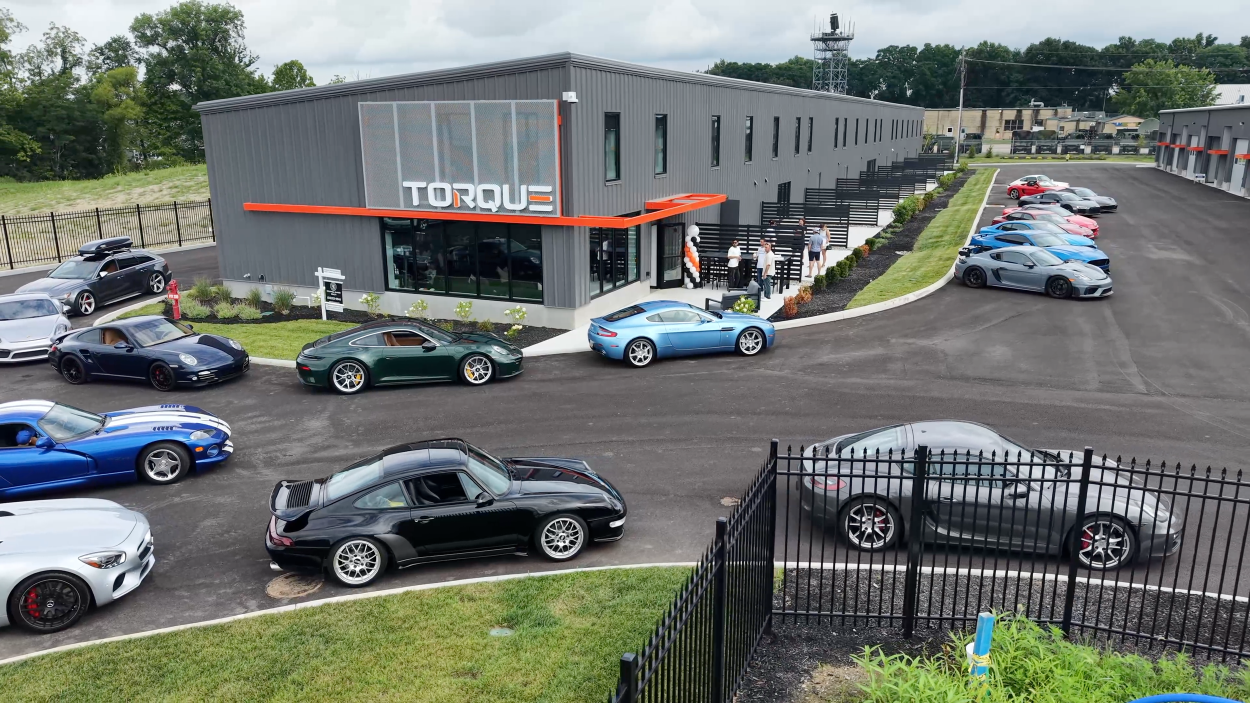 A parking lot filled with luxury cars in front of a modern building with the sign 'TORQUE' at the entrance. Several people are gathered outside near the building.