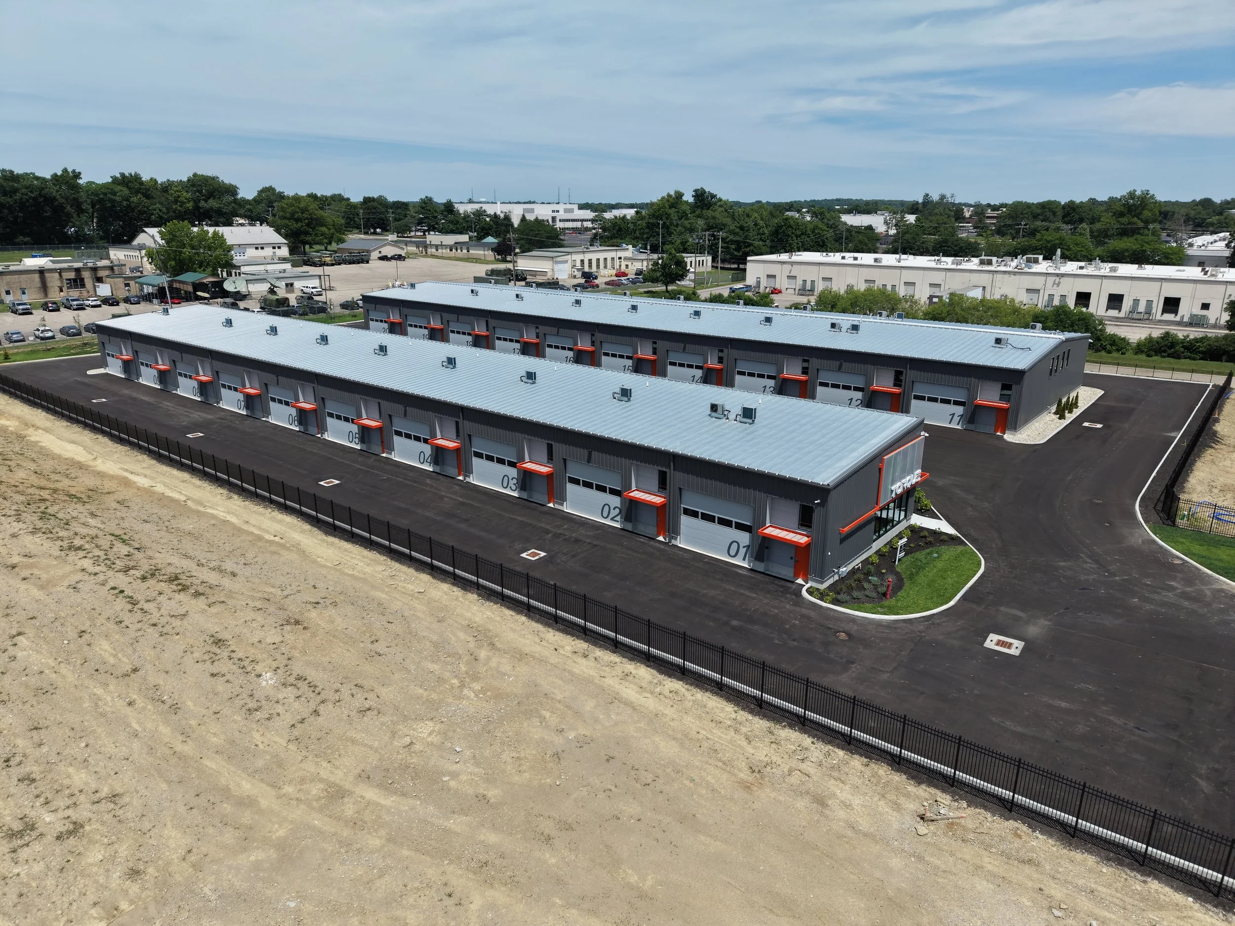 Aerial view of a large warehouse or industrial building with a gray roof and rows of numbered garage doors, surrounded by parking lots and other industrial buildings, with trees and a partly cloudy sky in the background.