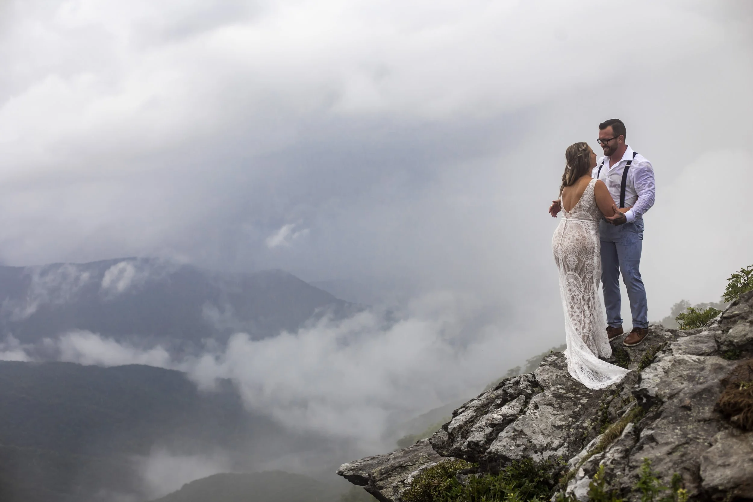 image of a woman in crocheted white gown on a mountaintop, embracing a man. mountains and clouds fill the background on a dark day