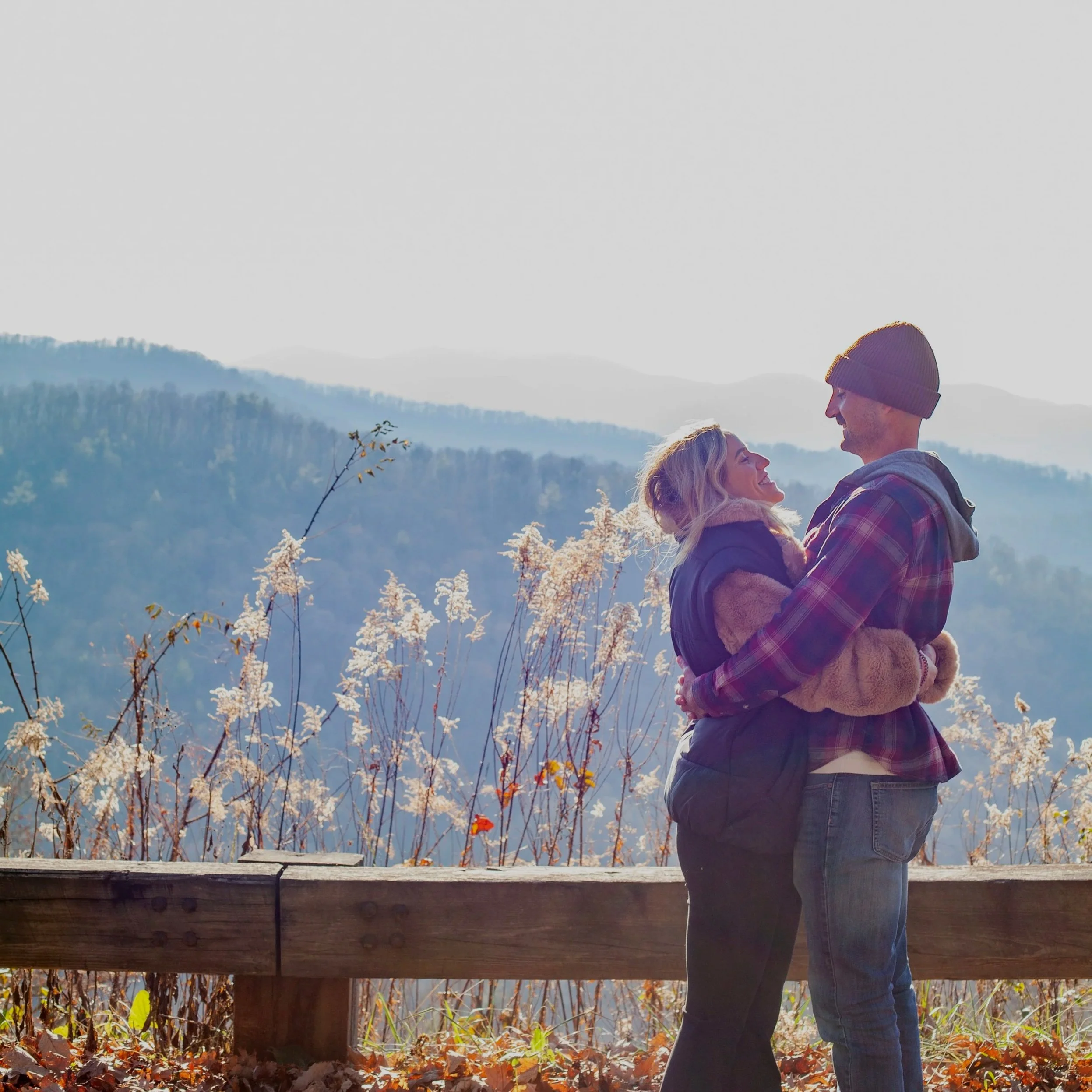 A couple embracing outdoors on a wooden deck with a scenic mountainside and lake in the background. The woman has blonde hair and is wearing a purple jacket, while the man is wearing a gray hoodie and a beanie. The scene is surrounded by fall foliage