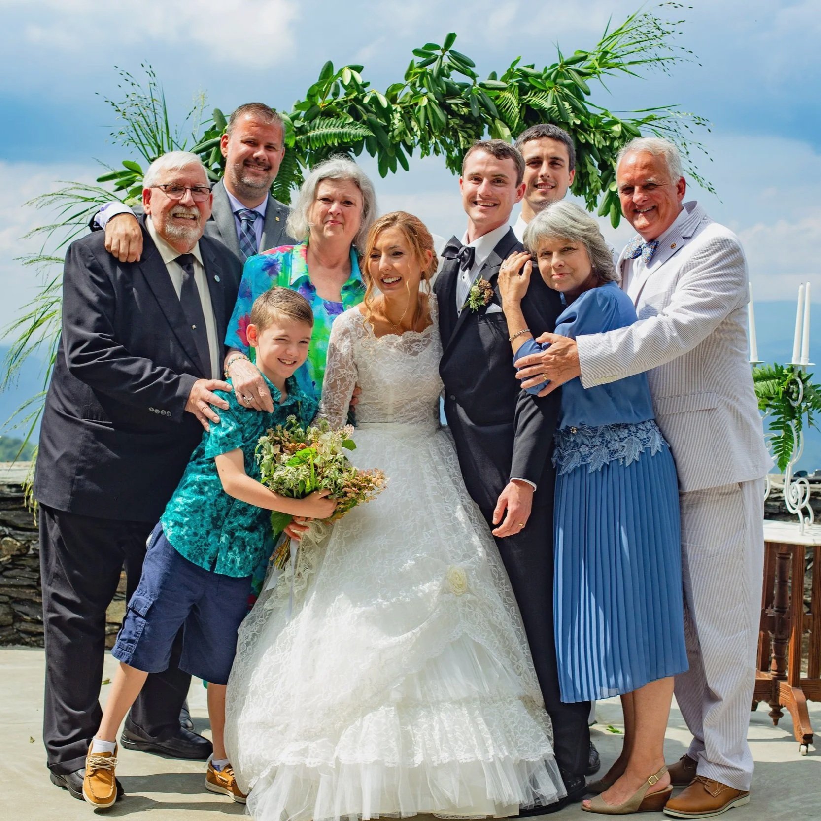 A group of ten people, including a bride and groom, posing outdoors under a leafy arch with a sky background. The bride is wearing a white lace wedding dress holding a bouquet, and the groom is in a black tuxedo and bow tie. The others include older 
