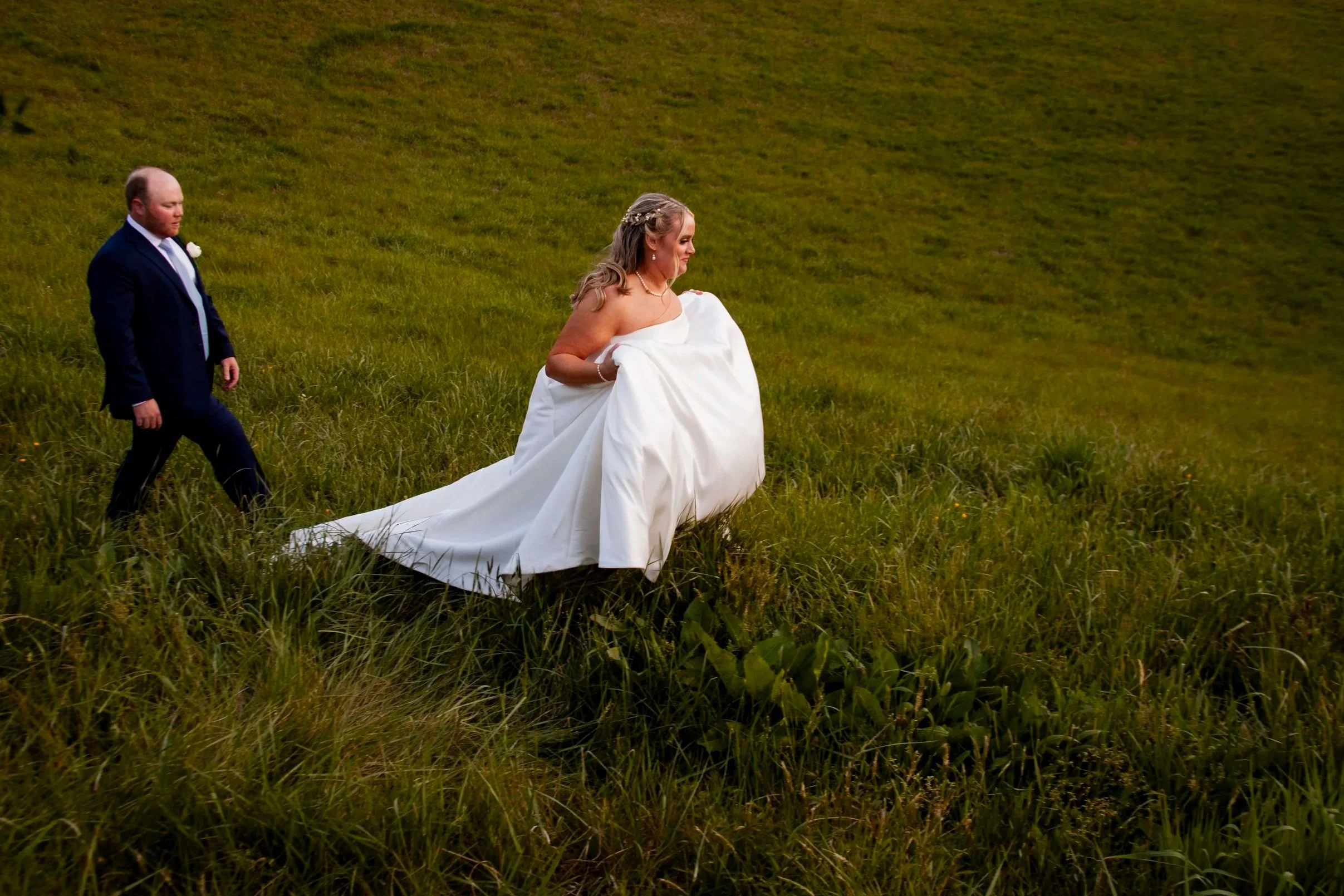 A bride in a wedding dress carrying her gown and walking through a grassy field, with a groom in a dark suit walking nearby.