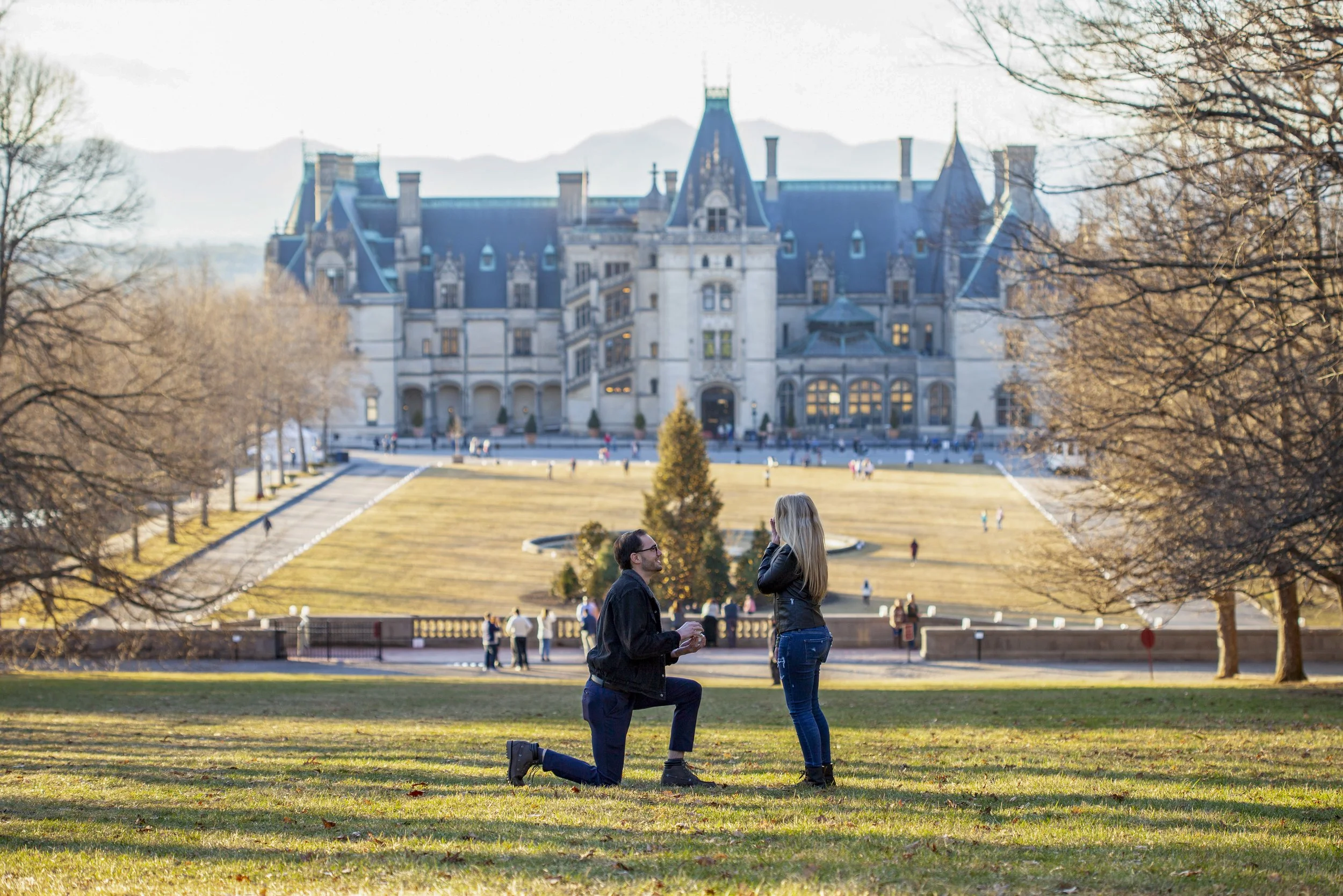 A couple is kneeling on the grass in front of a woman, facing each other, with a historic castle-like building in the background. Trees surround the scene, and other people are visible walking or sitting on the lawn.
