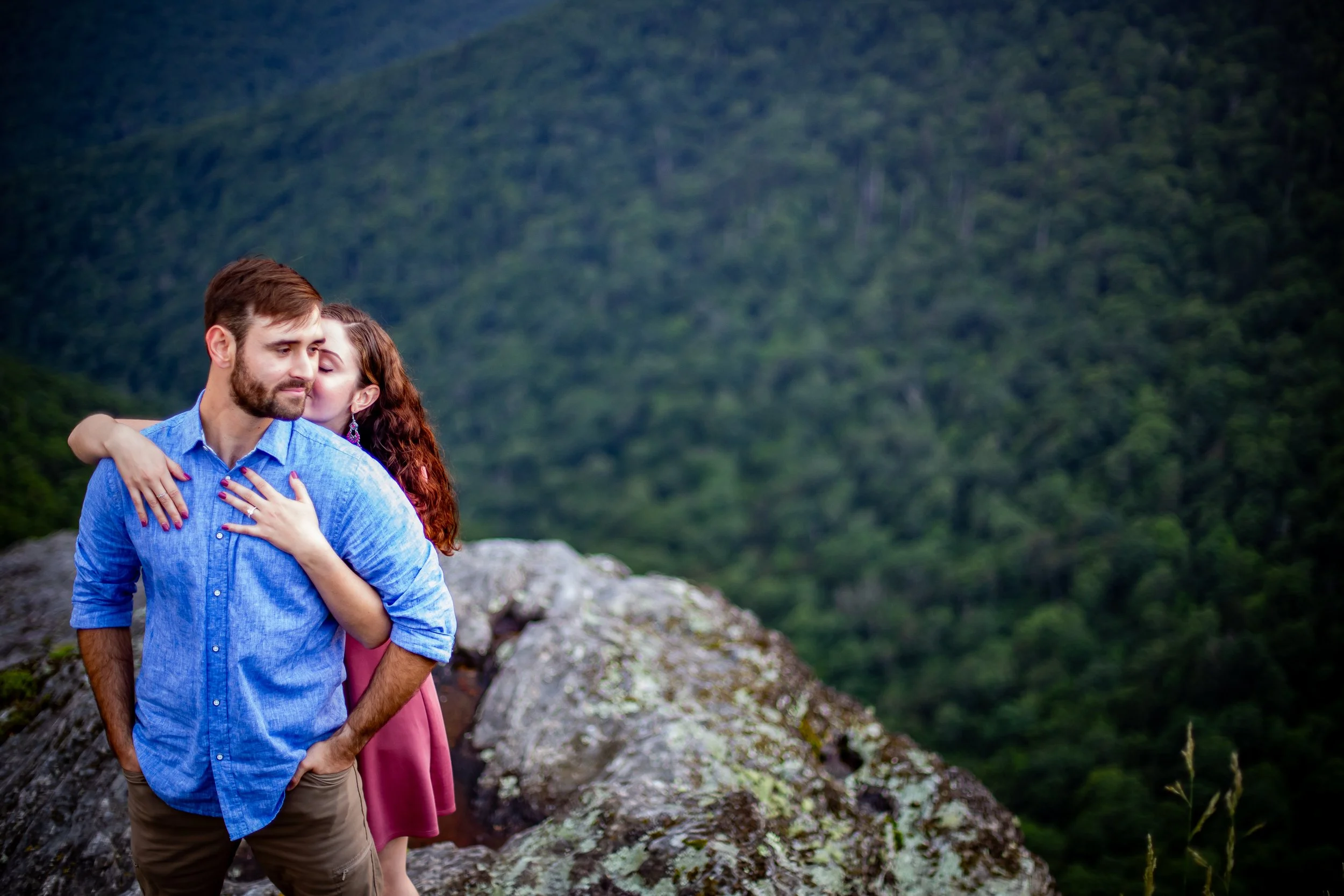 A couple standing on a rocky terrain outdoors, with a green forest in the background. The woman is kissing the man's cheek, and he is looking ahead with a slight smile.