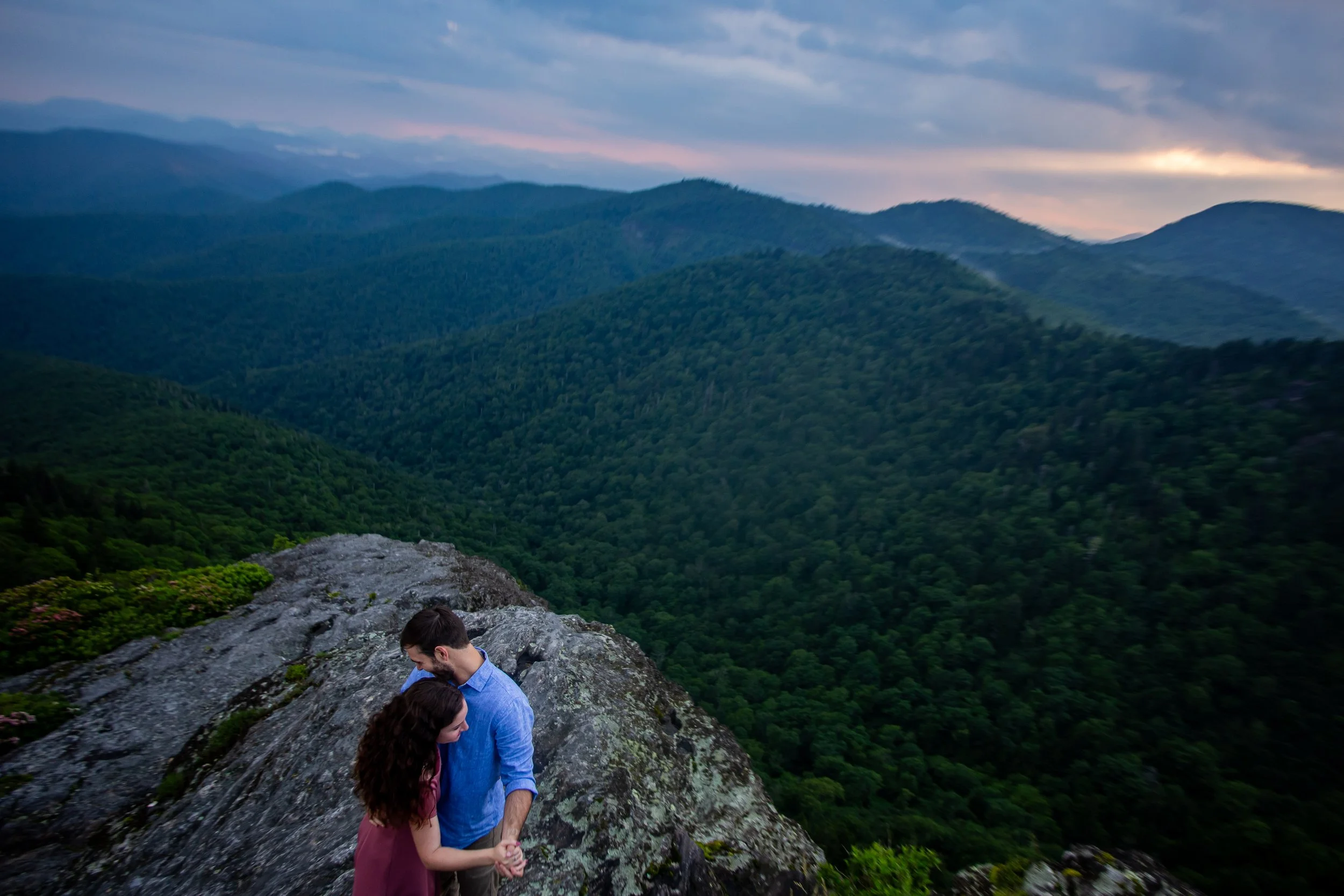 A couple holding hands and embracing on rocky terrain overlooking a lush green valley with mountain ranges and a cloudy sky at sunset.