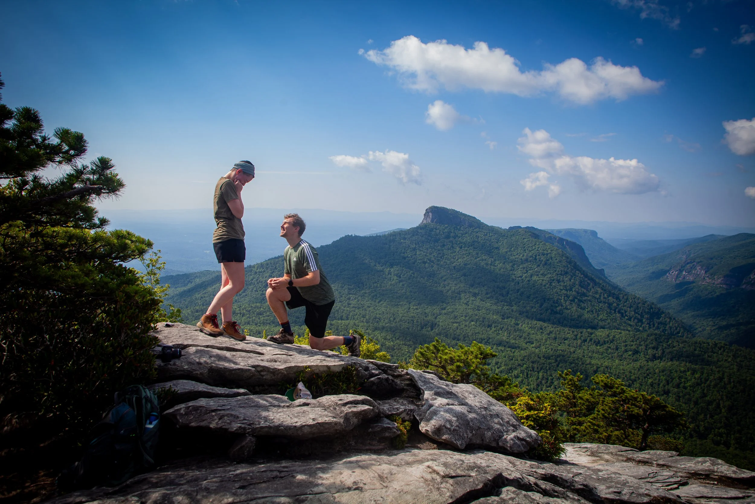 Two people are on a mountain peak with a scenic view of green forested mountains and a partly cloudy sky. One person is kneeling while the other is standing, with both appearing to be engaged in a romantic proposal.