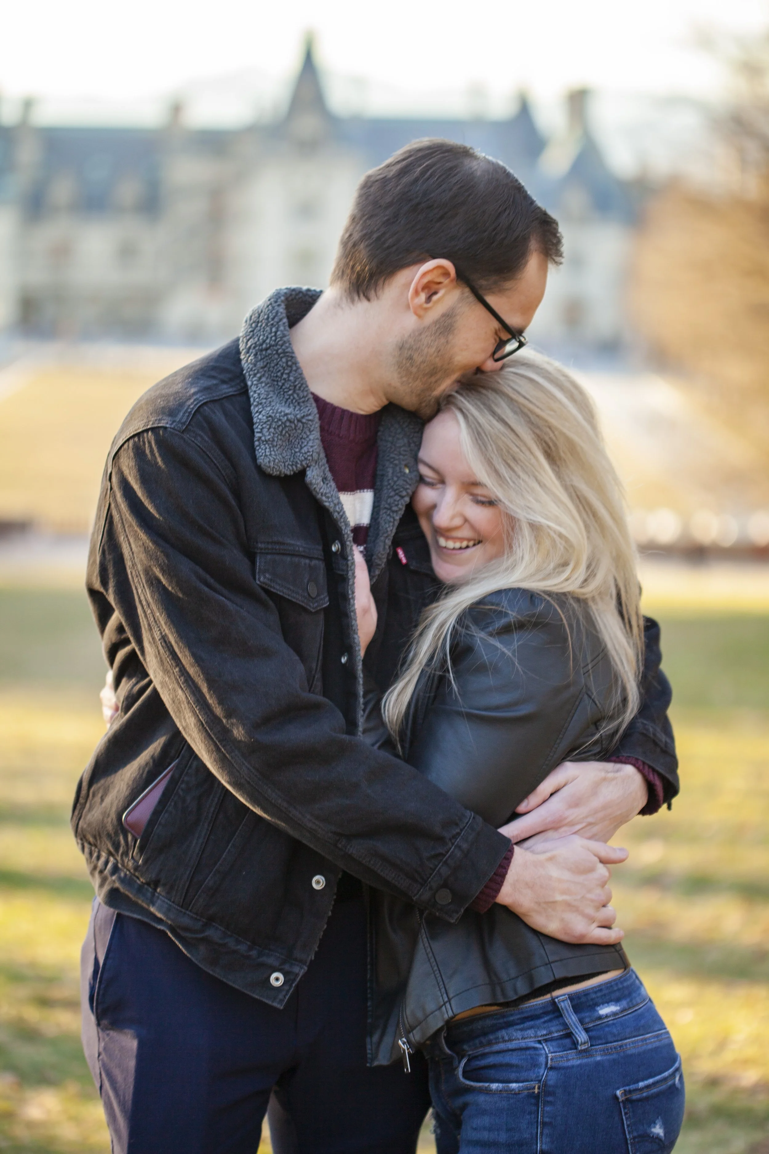 A man wearing glasses and a dark jacket is hugging a woman with blonde hair and a black leather jacket outdoors, with a blurry historic building in the background, during daytime.
