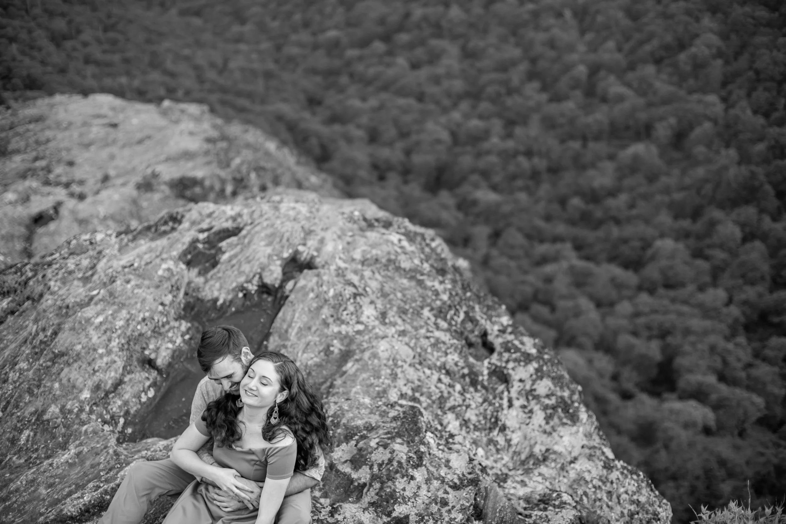 A young couple sitting and embracing on a rocky ledge in a forested area with rolling hills in the background.