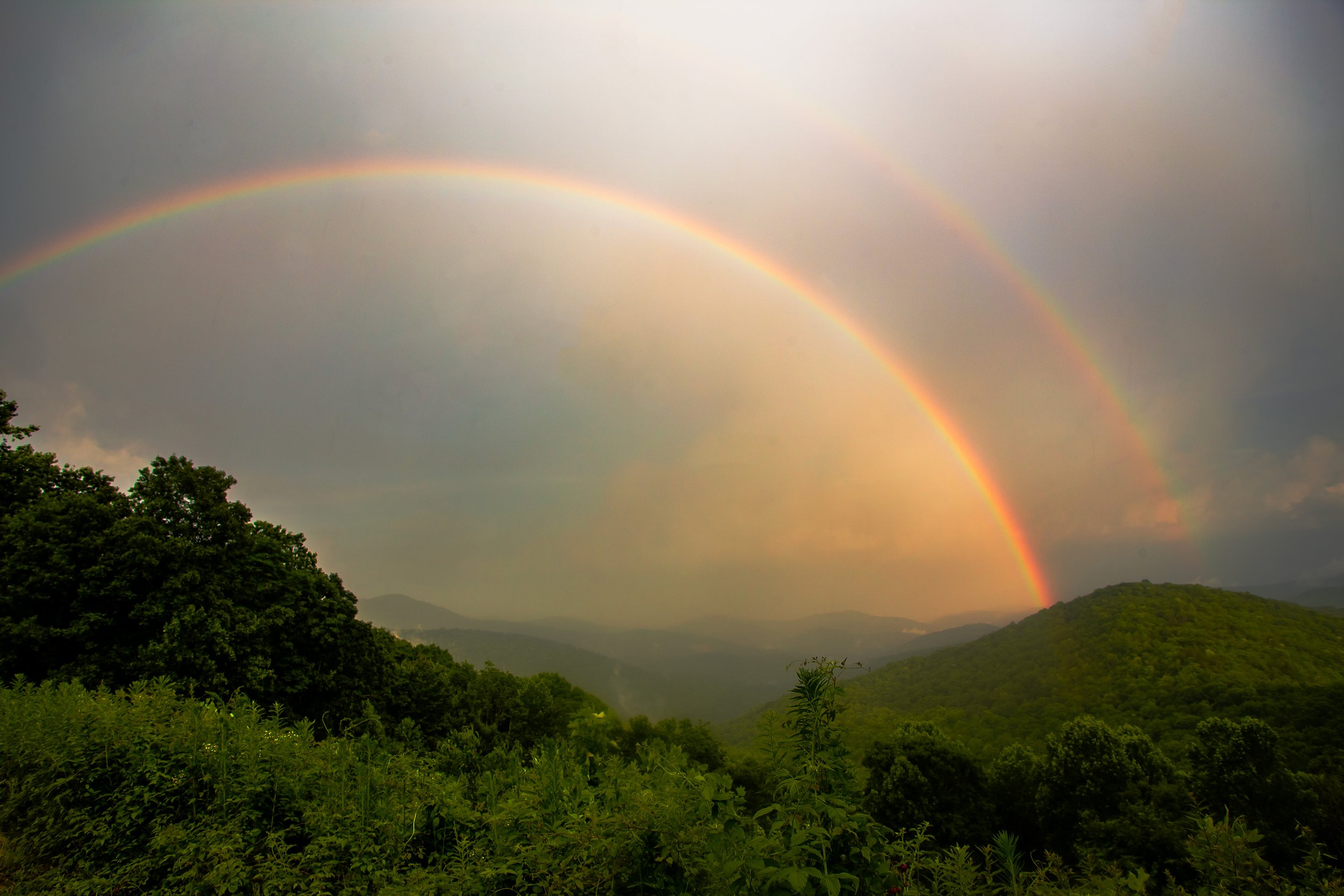 A double rainbow over a lush green landscape with trees, rolling hills, and mountains in the background, under a cloudy sky.