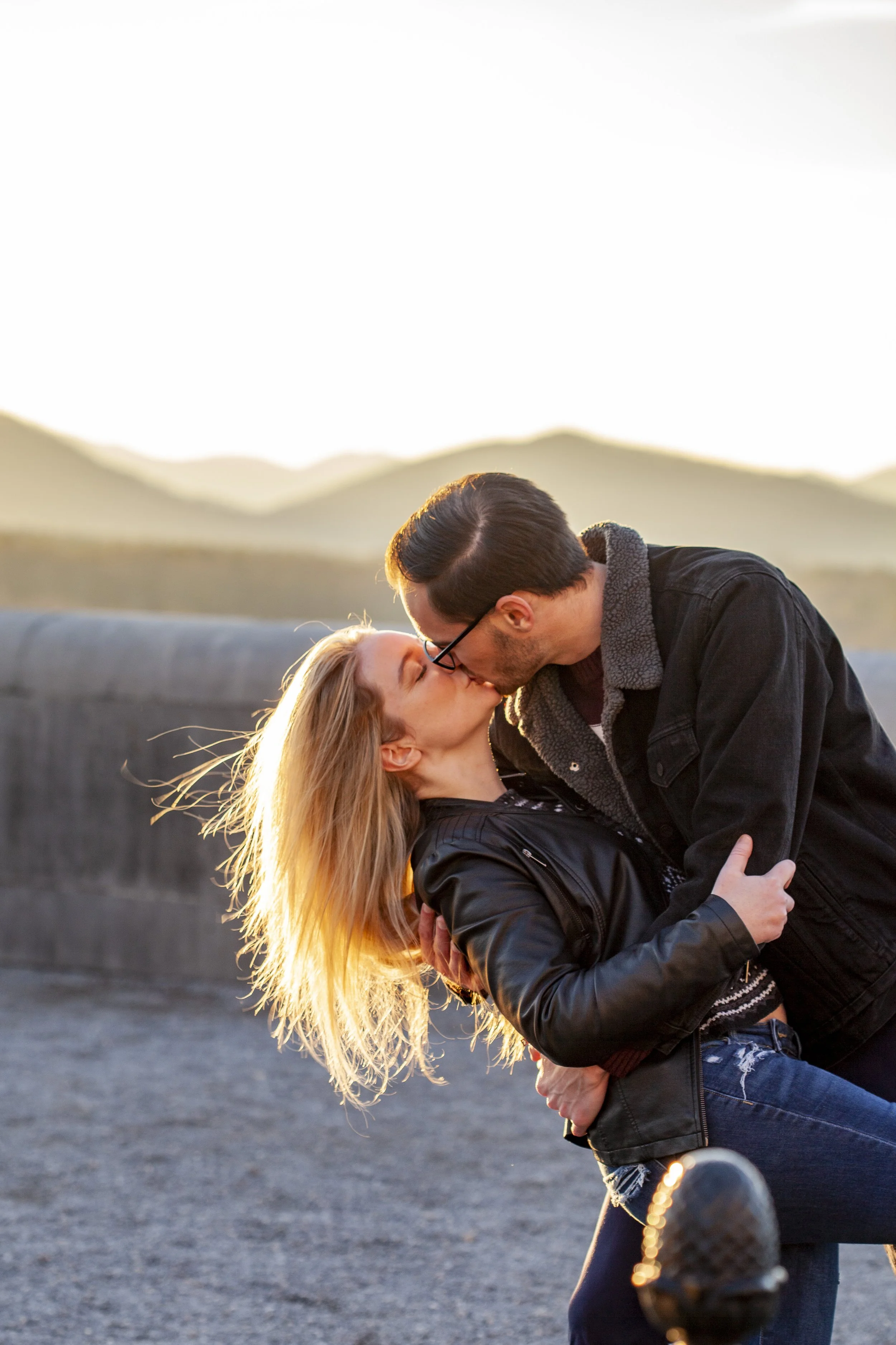 A couple sharing a kiss outdoors during sunset, with the man bending down and the woman leaning back, hair illuminated by the sunlight, both wearing jackets.