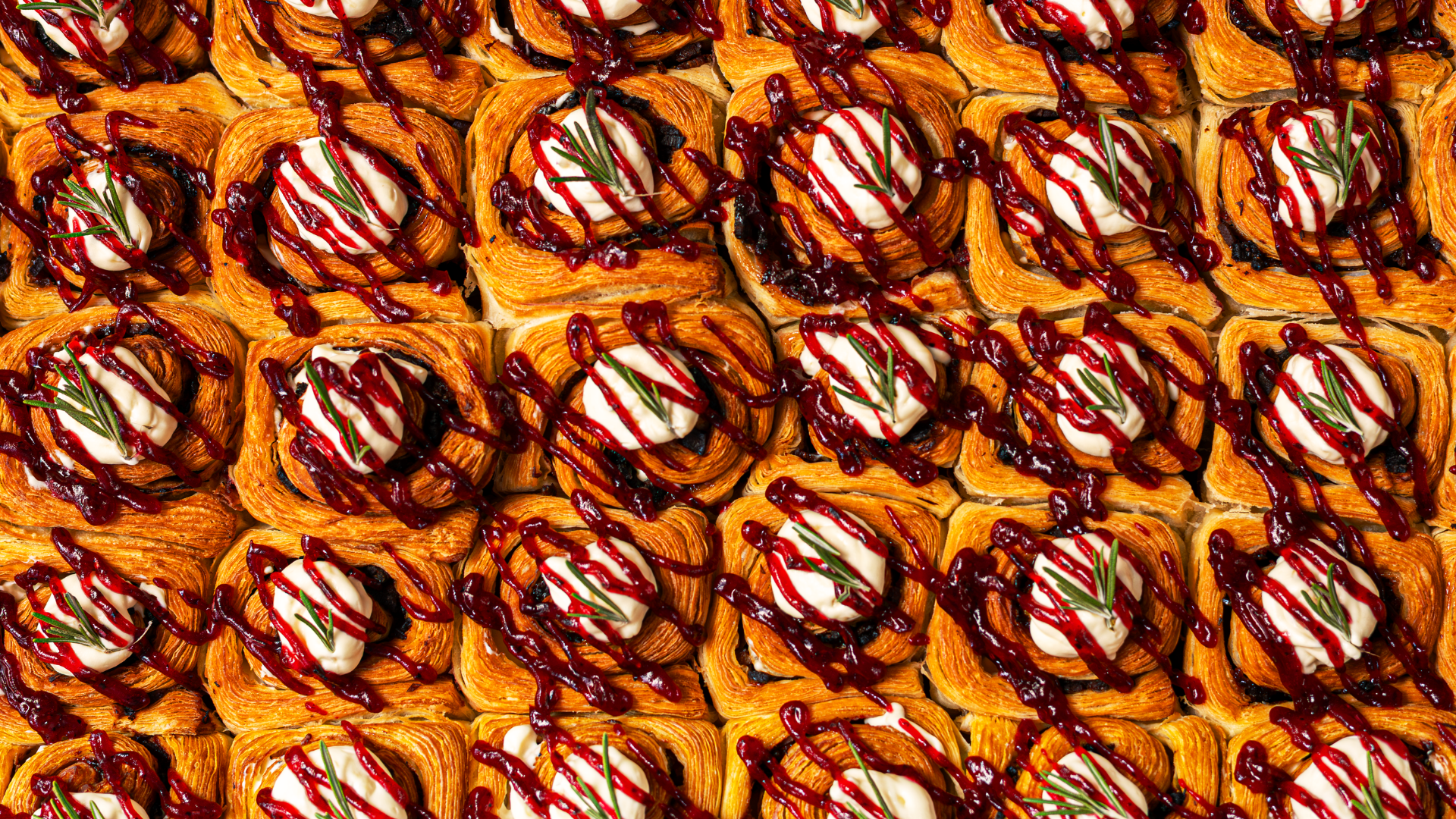 A close-up of multiple mini savory pastries topped with a dollop of cream, garnished with rosemary sprigs, and drizzled with red berry sauce.
