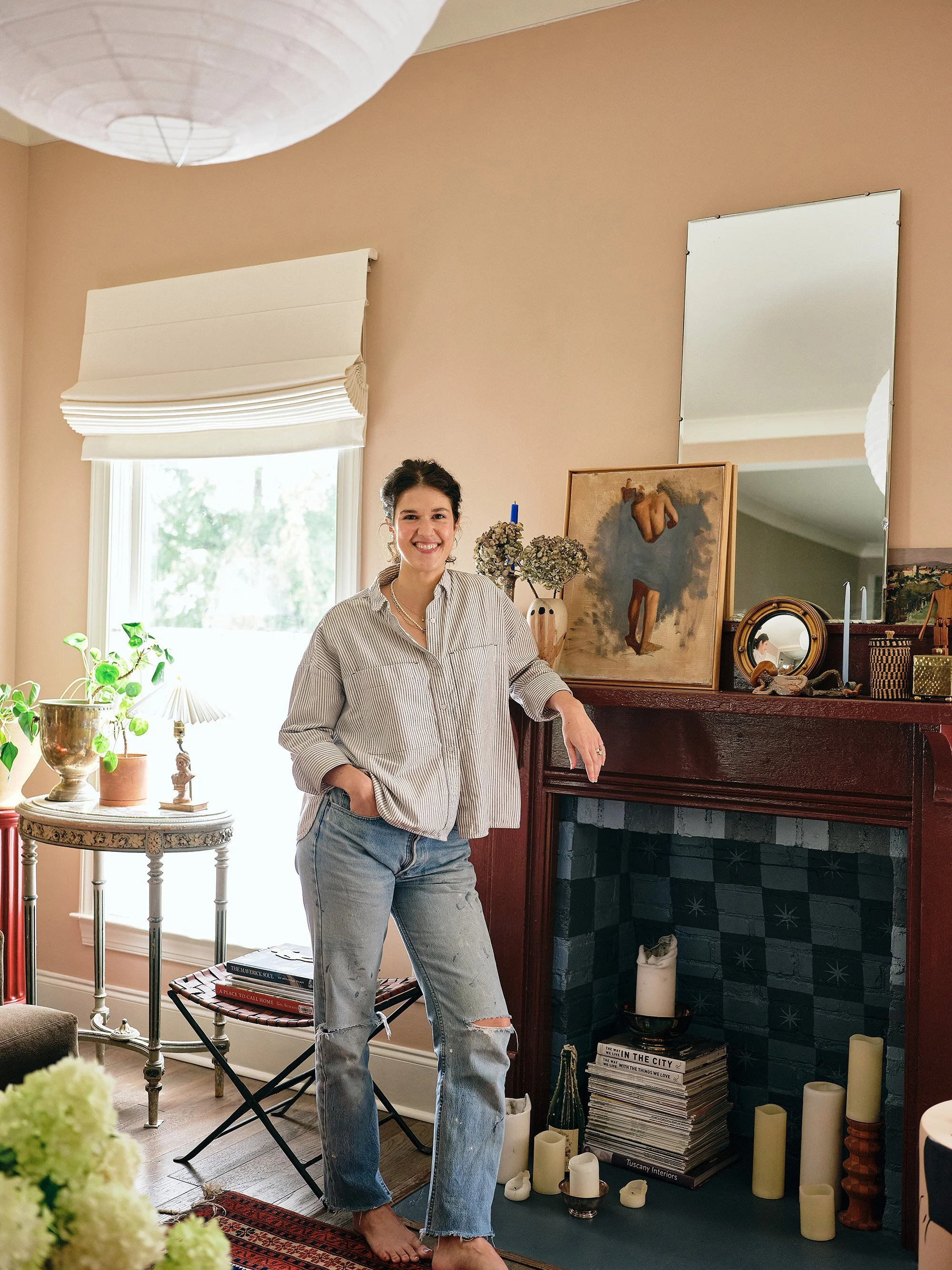 A young woman with dark hair, wearing a striped shirt and ripped jeans, smiling and standing by a fireplace in a cozy room with decorative objects, plants, and artwork.