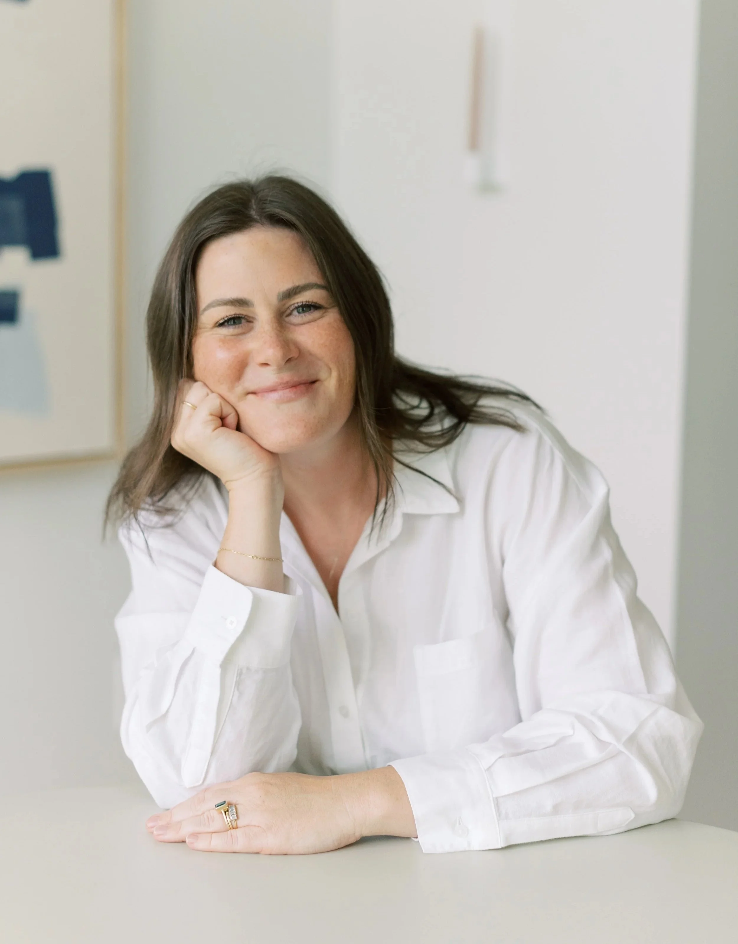 A woman with long brown hair and blue eyes, wearing a white button-up shirt, resting her chin on her hand, smiling, with rings on her fingers, sitting at a light gray table in a bright room.