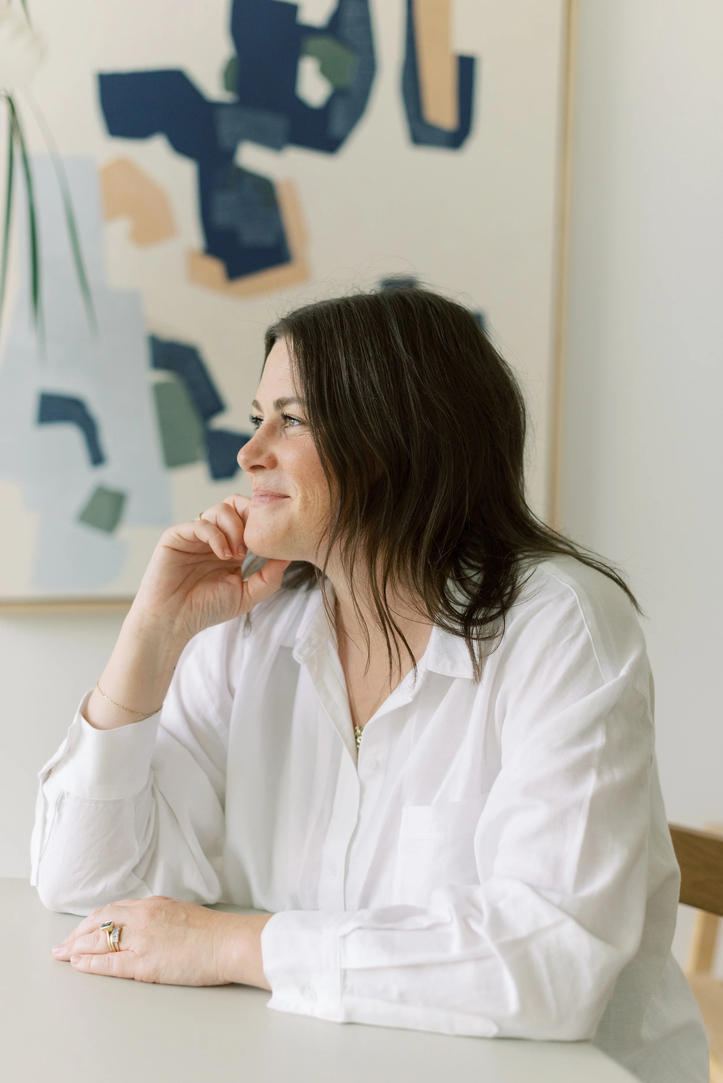 A woman with dark brown hair wearing a white lab coat, sitting at a table with her chin resting on her hand, in an indoor setting with blurred abstract artwork on the wall behind her.