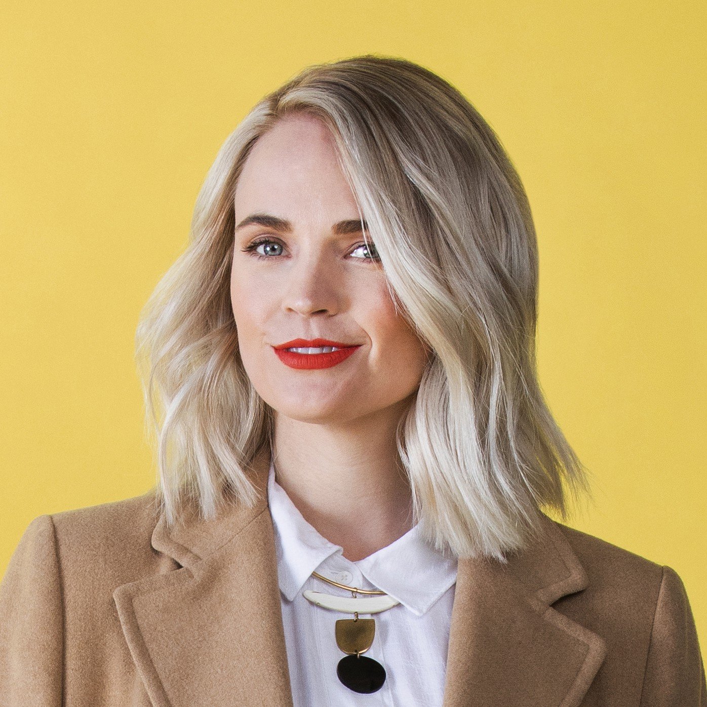 A woman with shoulder-length blonde hair, red lipstick, wearing a white blouse, a beige blazer, and a necklace with large circular pendants, standing against a yellow background.