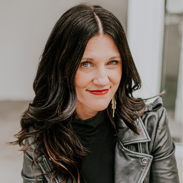 A woman with long black wavy hair, wearing a black shirt and a black leather jacket, smiling at the camera indoors.