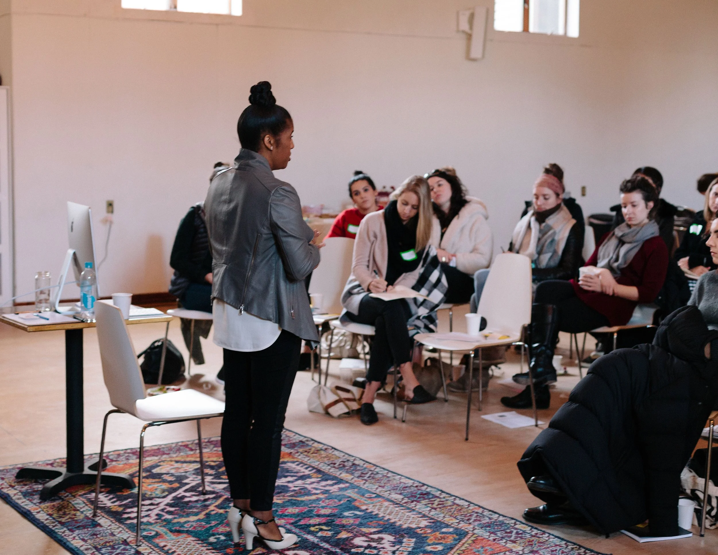 A woman standing in front of a group of seated women during a presentation or workshop in a well-lit room with high windows and a patterned rug.