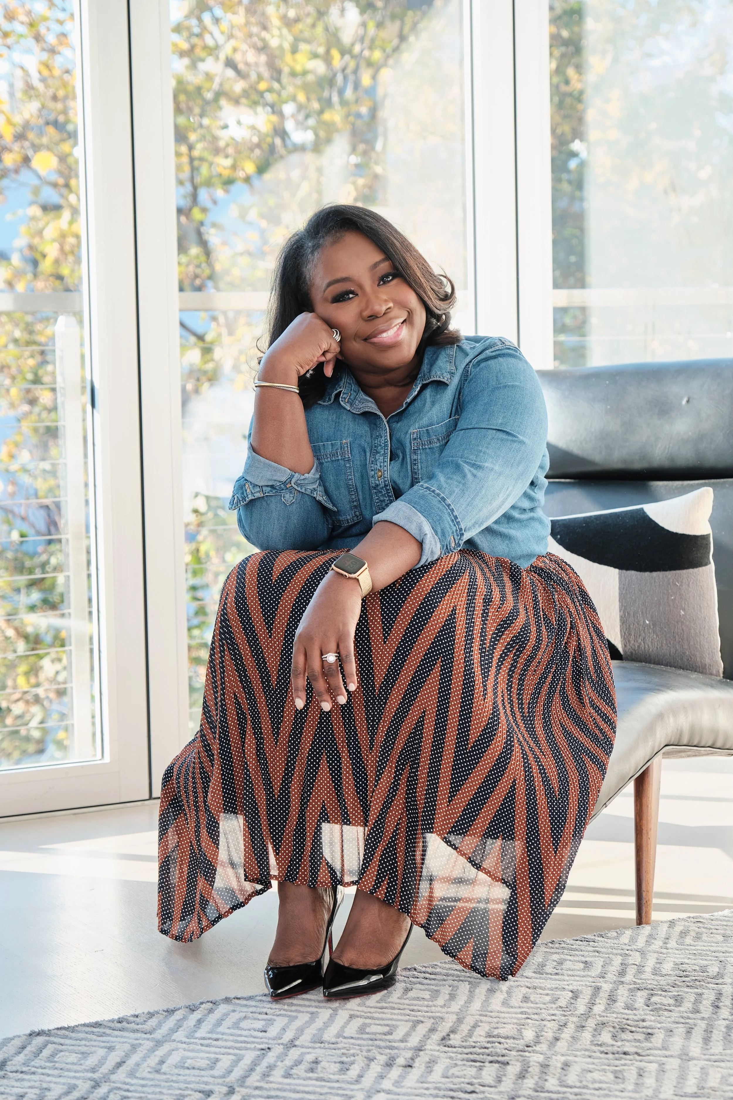 A woman sitting in a chair with a big smile, wearing a denim shirt, a patterned skirt, high heels, a watch, and rings, in a room with large windows showing trees outside.