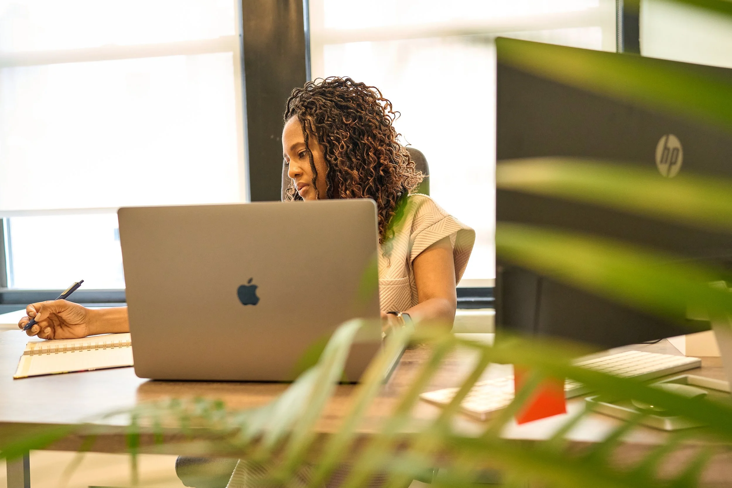Woman with curly hair working at a desk with a laptop, notebook, and pen in an office setting, with green plants in foreground and bright windows in background.