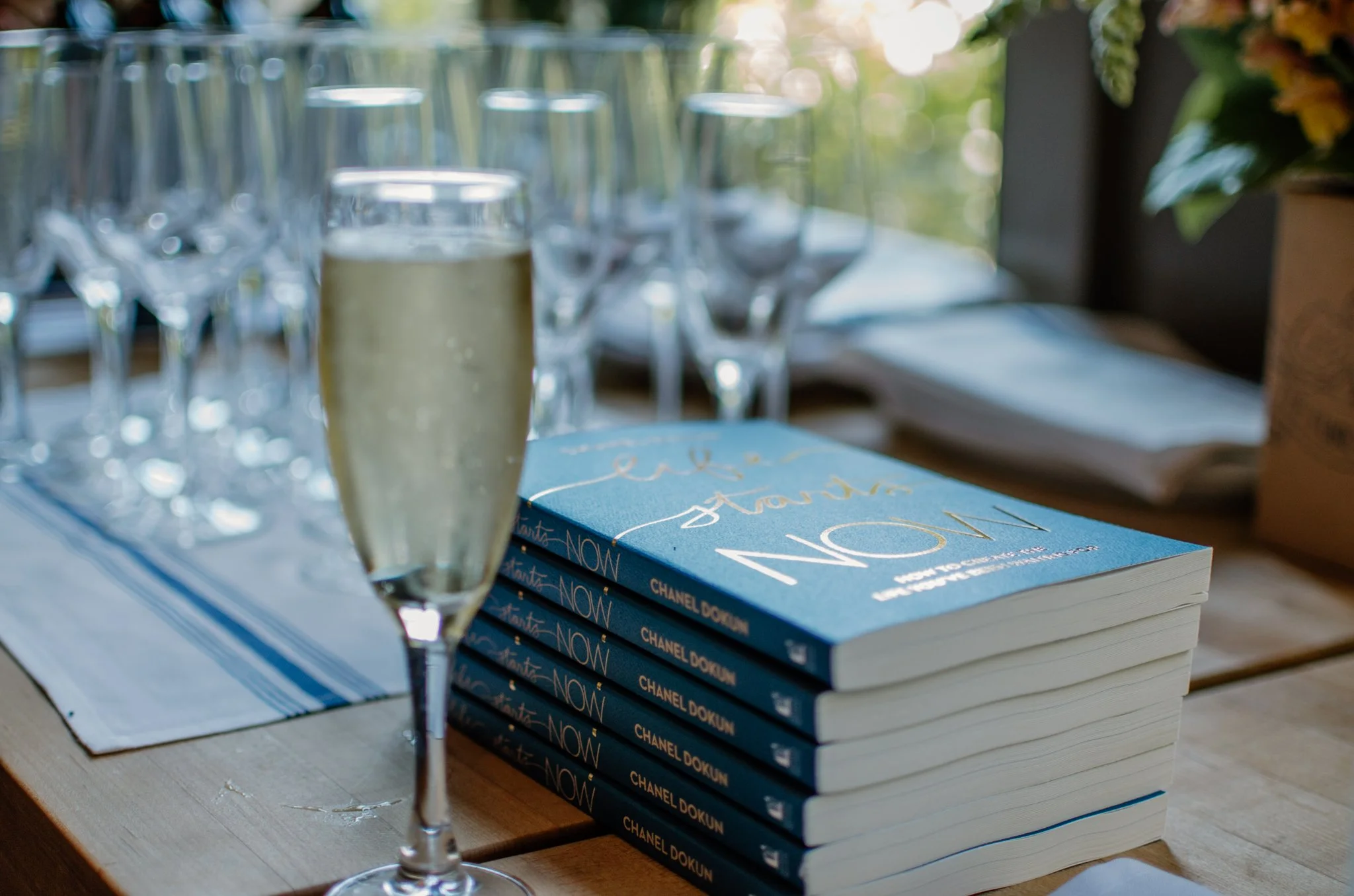 A filled champagne flute near a stack of blue books titled 'Chanel Dokun' on a wooden table, with multiple empty glasses and a blurred window in the background.