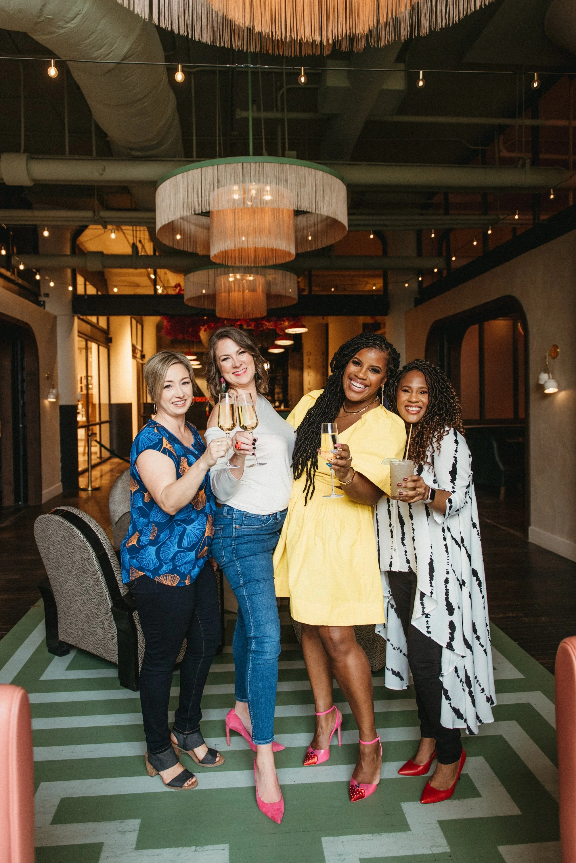 Four women at a social gathering holding glasses of champagne and a cocktail, standing on a green and white striped floor inside a modern restaurant or bar with warm lighting and hanging ceiling fixtures.