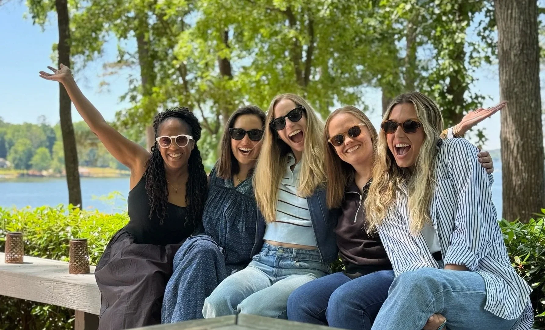 Group of five women sitting outdoors near a body of water, smiling, wearing sunglasses, with trees and blue sky in the background.