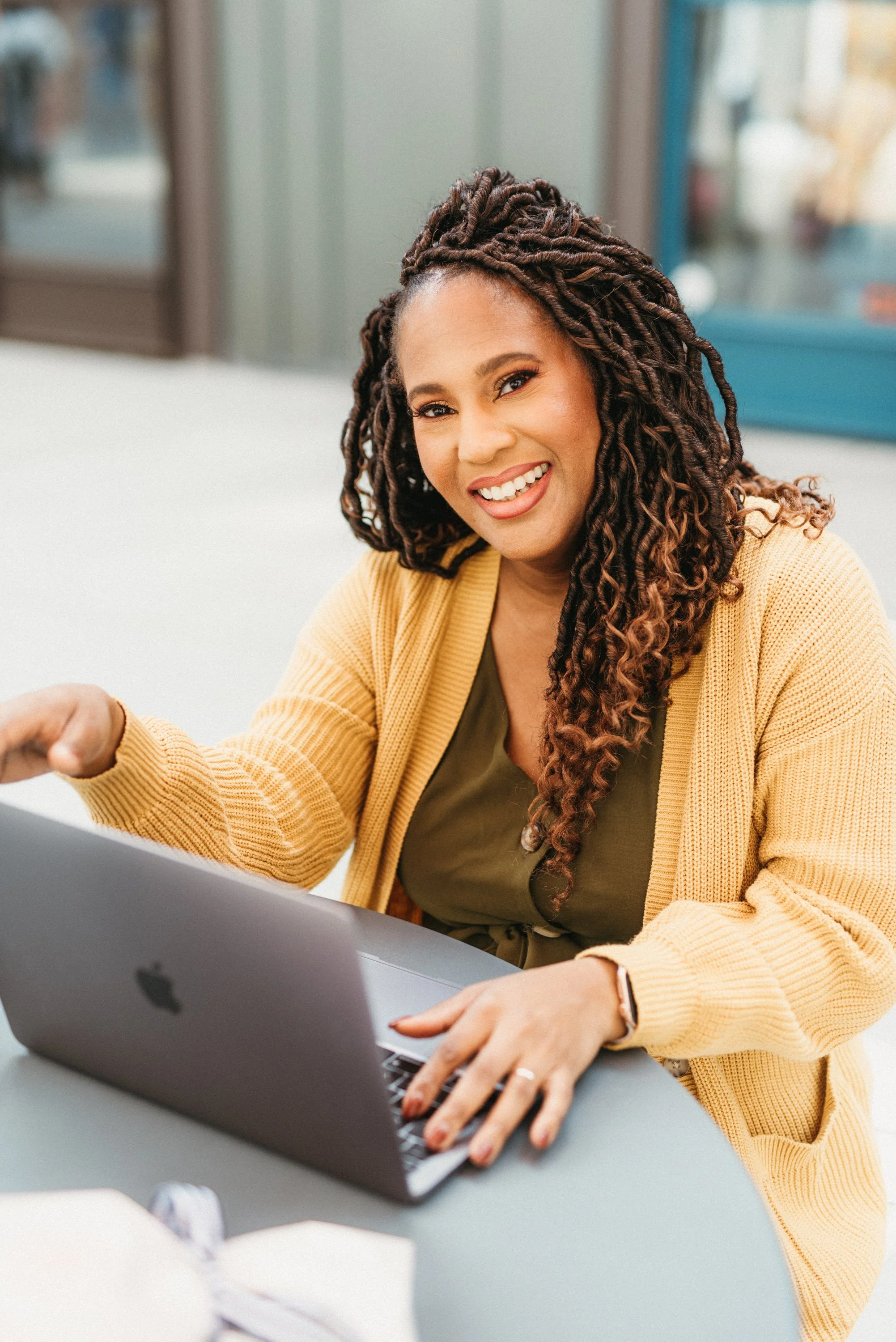 A woman with long, curly hair and a yellow cardigan working on a silver laptop in an office setting, smiling.