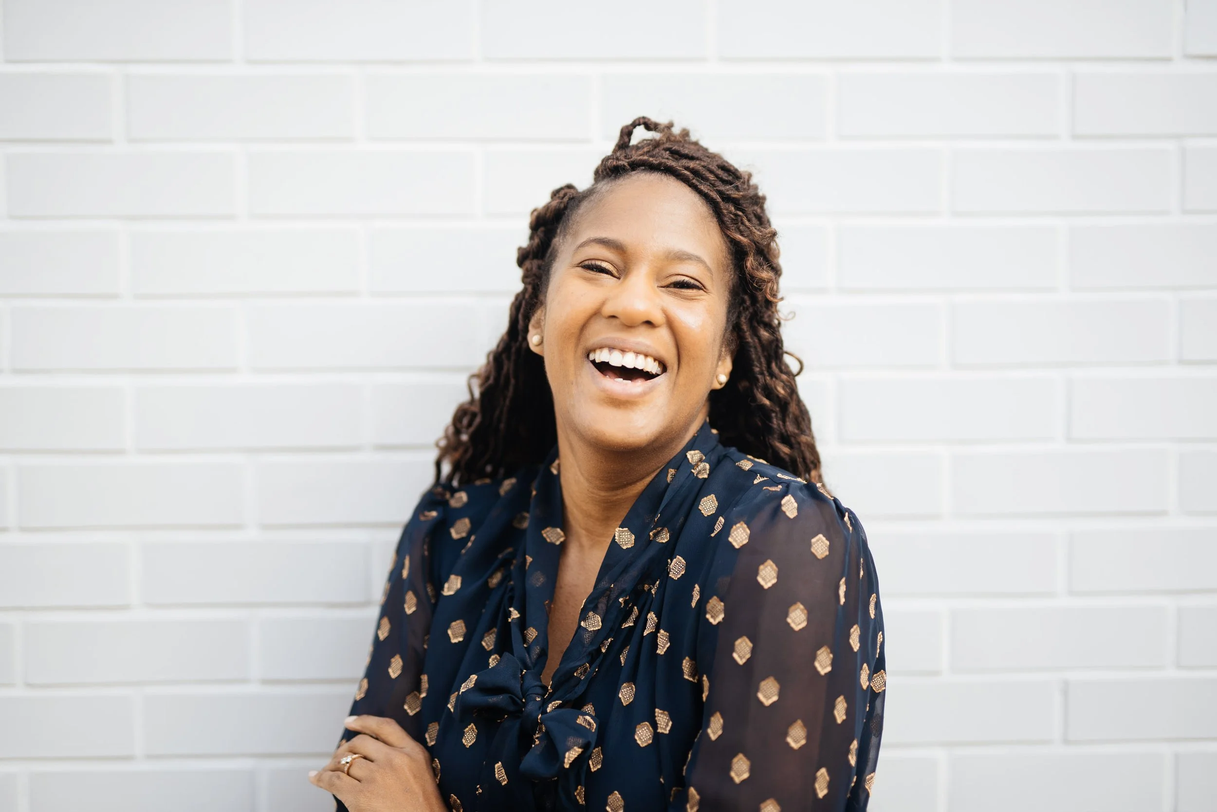 A woman with dreadlocks smiling and laughing, wearing a dark blue patterned blouse, standing in front of a white brick wall.