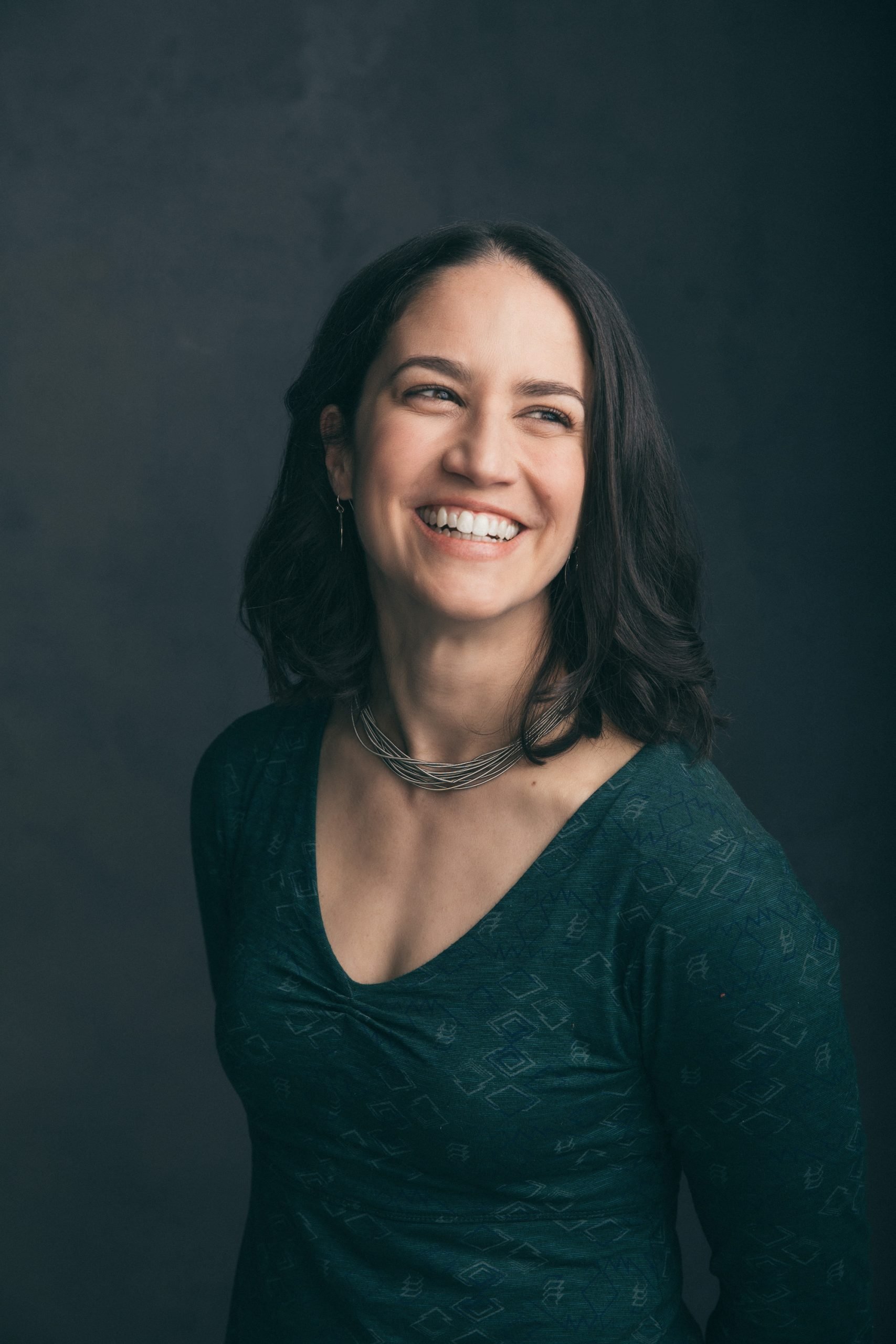 A woman with long dark hair smiling against a dark background.