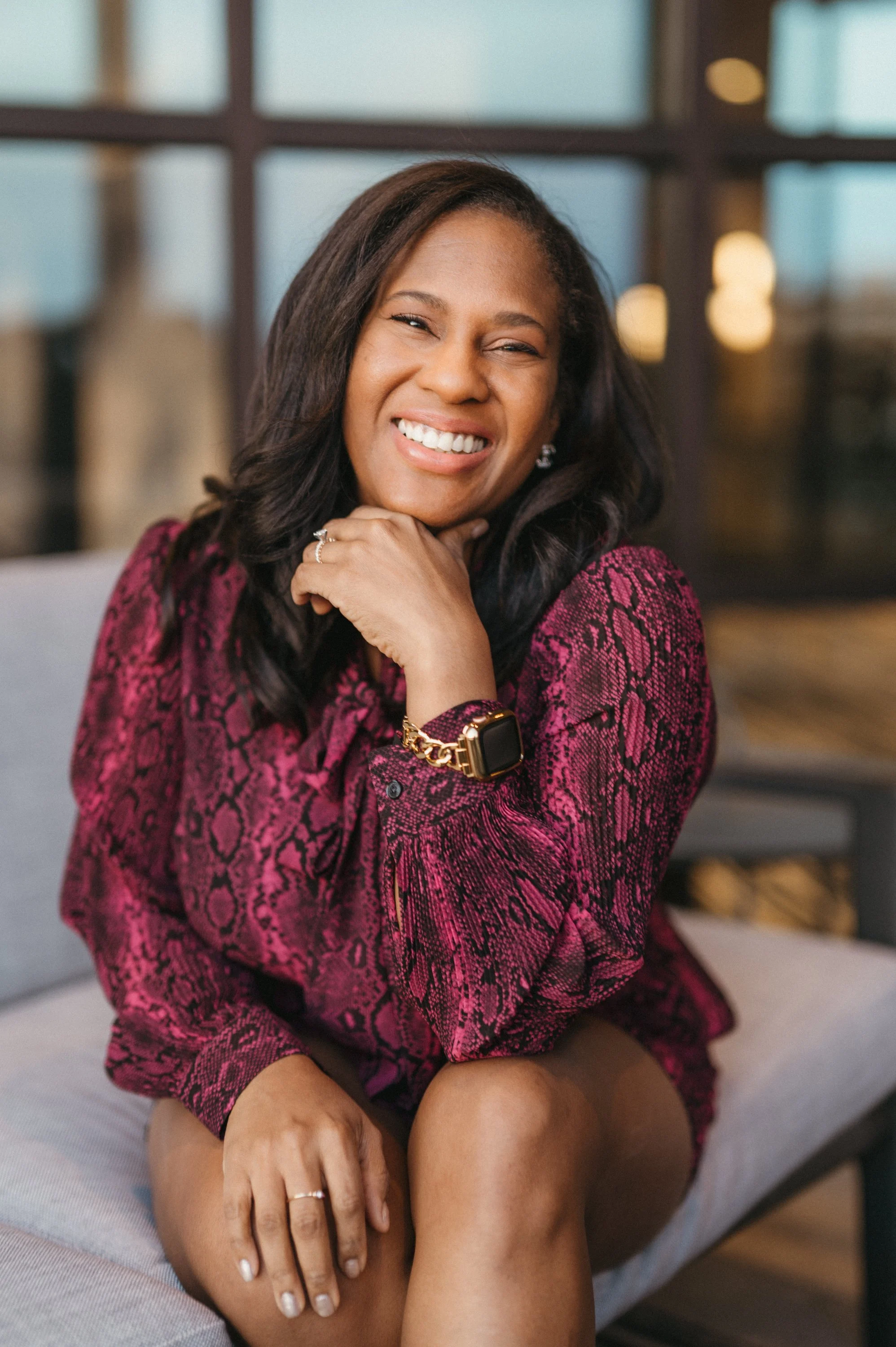 A woman smiling, wearing a red snake print jacket, sitting on a bed with one hand resting on her knee, in a warmly lit room with large windows in the background.