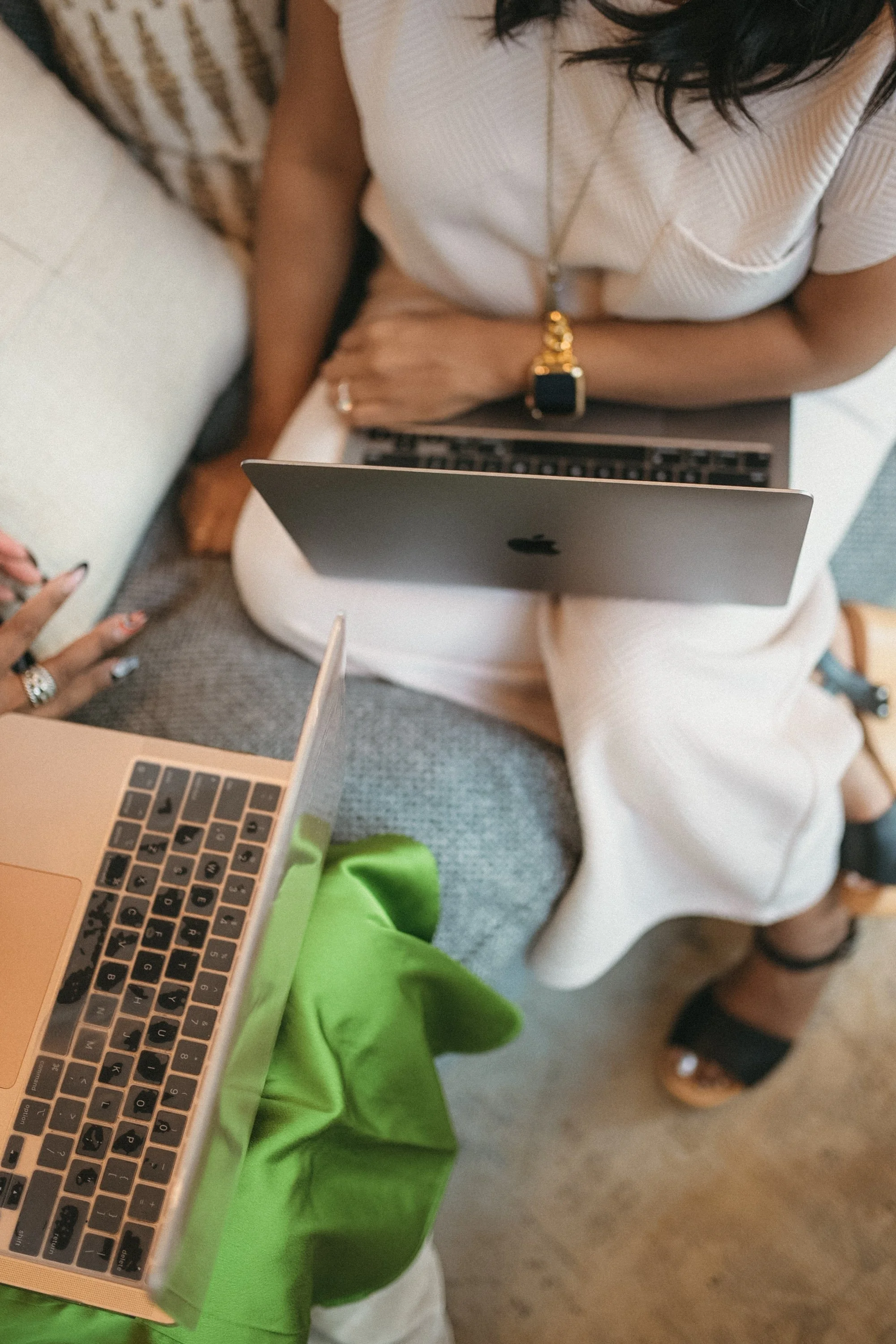 A person wearing a white shirt and white pants sitting with a laptop, a smartwatch on their wrist, and a gold necklace, with another person nearby and a green cloth in the foreground.