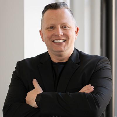 A smiling man with short gray hair, wearing a black blazer and black shirt, standing indoors near a window.
