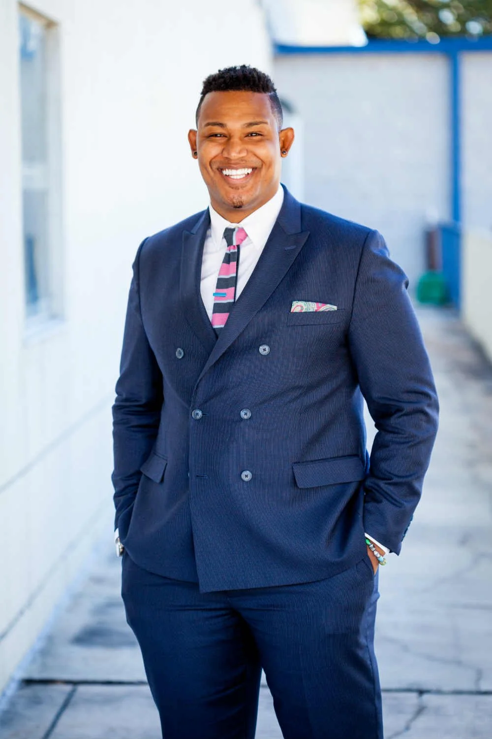 A man in a navy double-breasted suit, white shirt, and a pink, navy, and white striped tie standing outdoors with a smile.