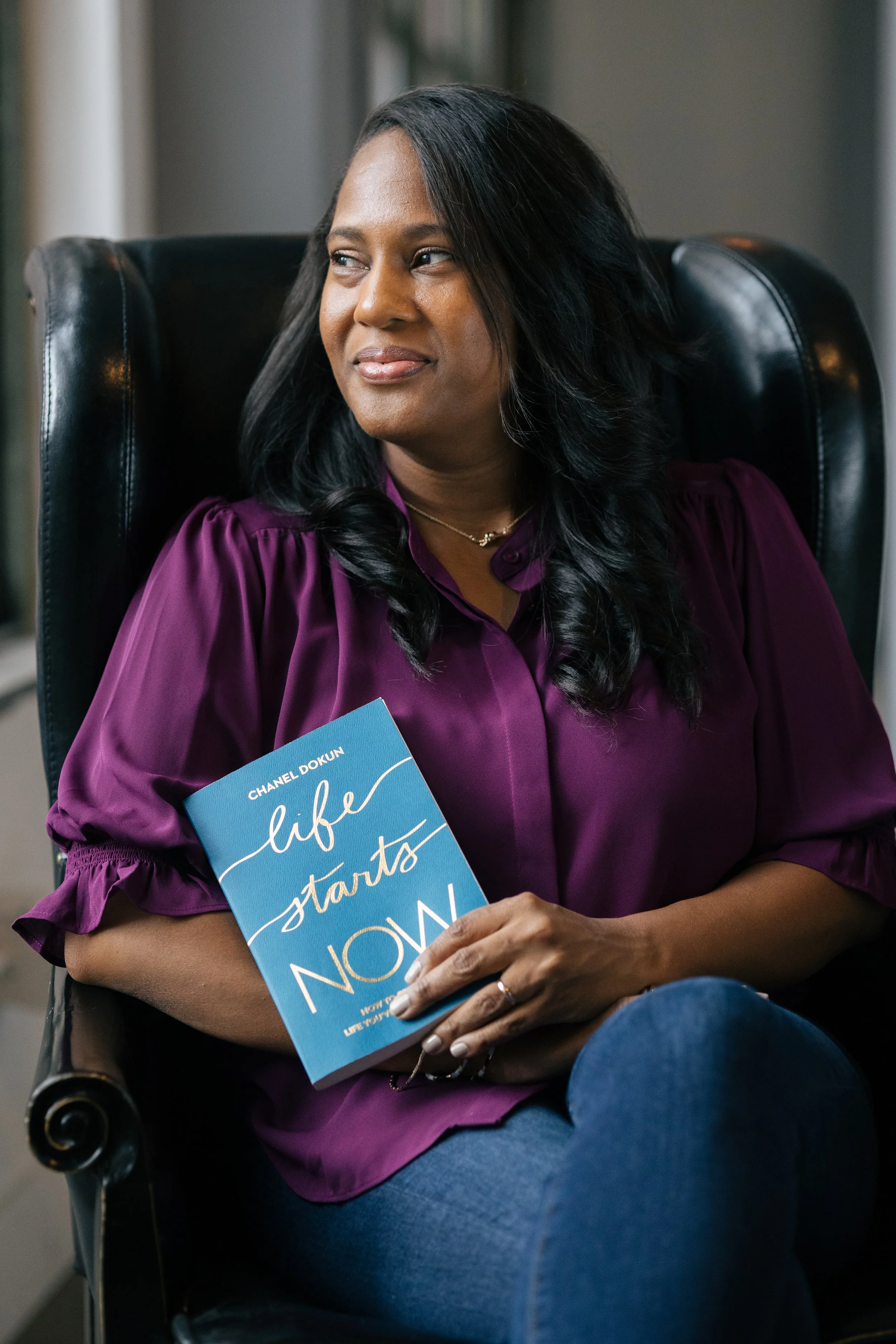 A woman with dark hair wearing a purple blouse, holding a blue book titled "Life Starts Now," sitting in a black leather chair.