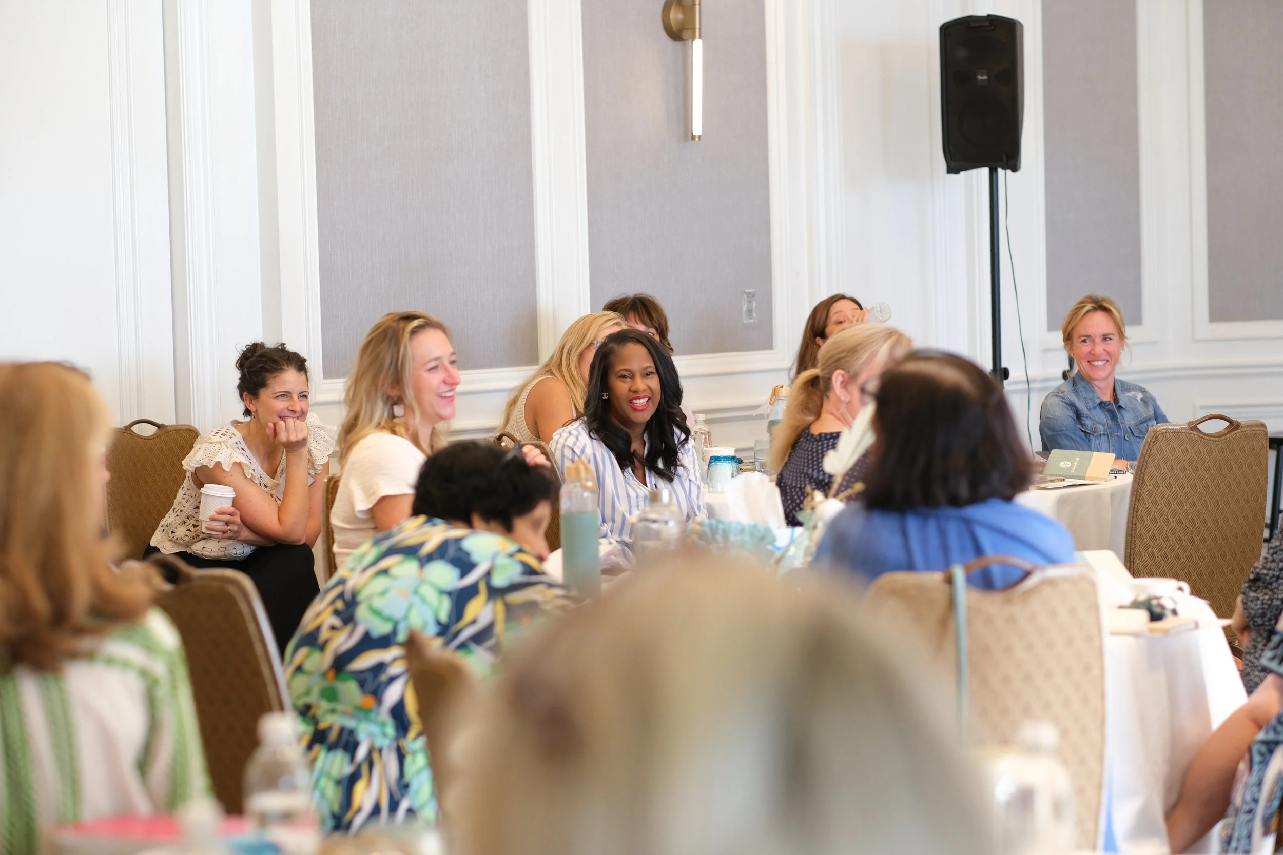 A group of women sitting at a conference table, smiling and engaging in a discussion during a meeting or seminar in a formal room with gray walls and a large speaker in the corner.