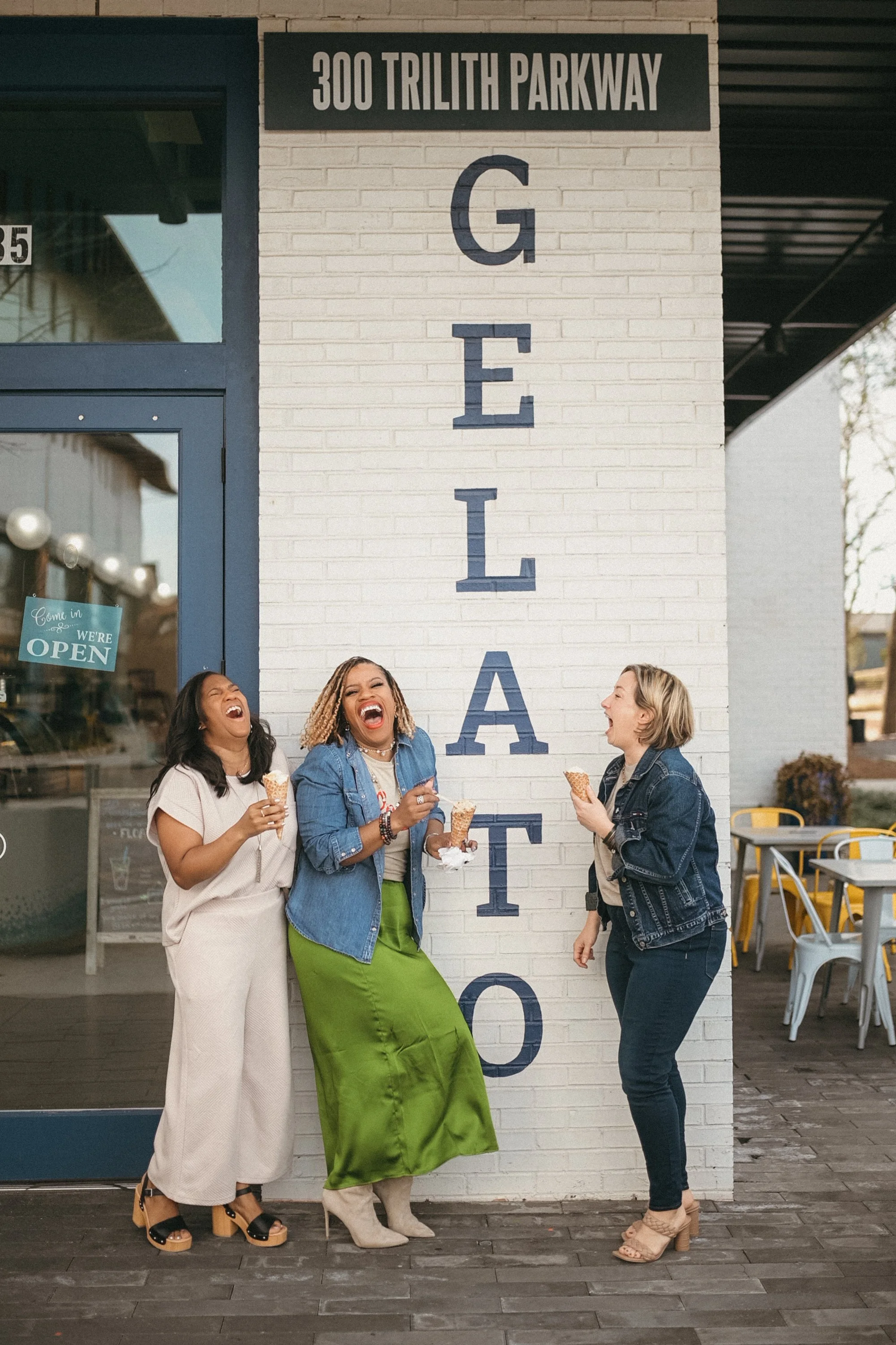 Four women laughing and talking outside a business with a sign on the wall that says '300 Trillith Parkway G E I A'. They are holding ice cream cones and are standing on a wooden deck, with outdoor seating and a large window with a sign that says 'We're Open' in the background.