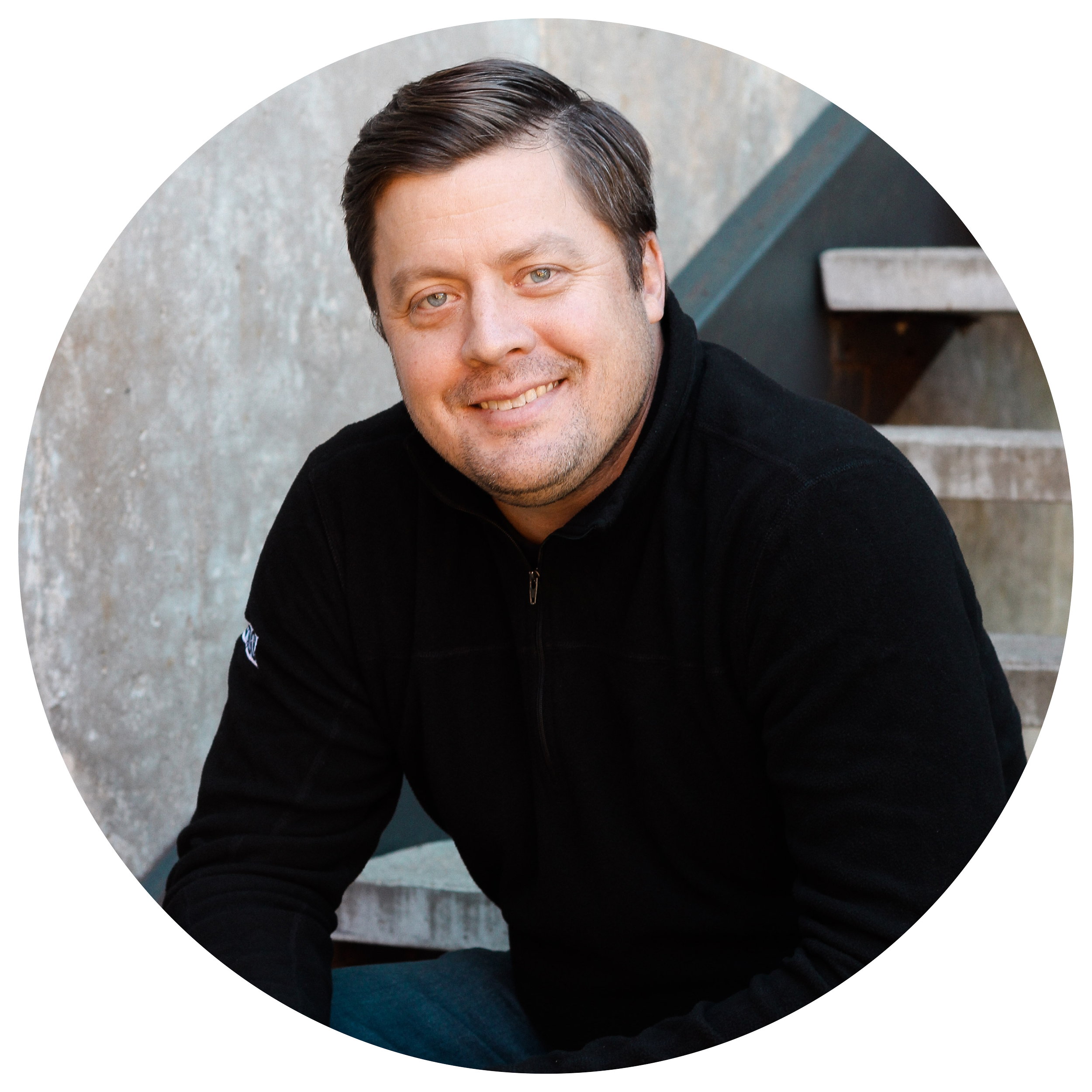 A smiling man with dark hair, wearing a black zip-up jacket, sitting outdoors near concrete stairs and a concrete wall.