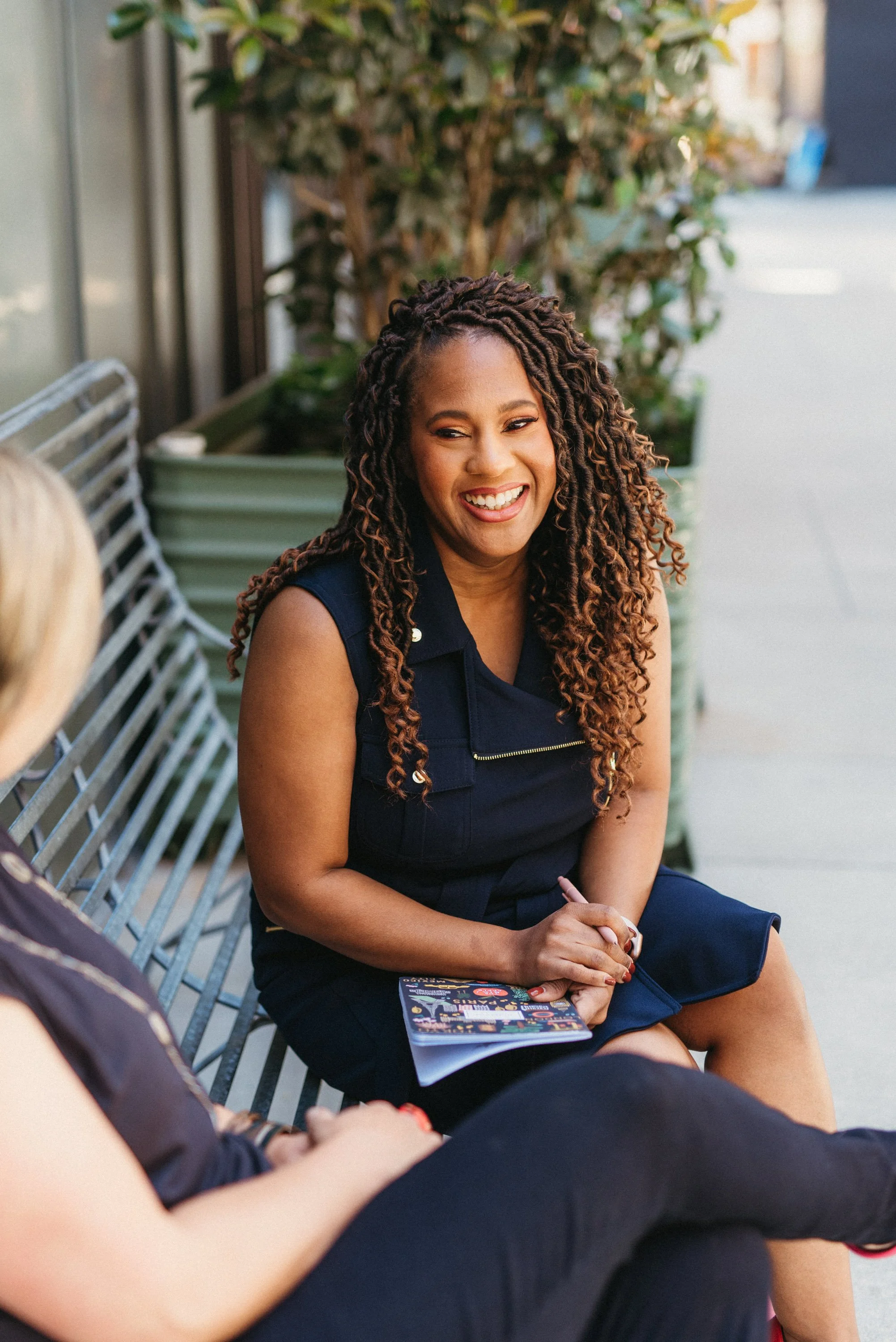 A woman with long, curly hair smiling and talking during a conversation outdoors, sitting on a metal bench with another person partially visible in the foreground, holding a colorful brochure.