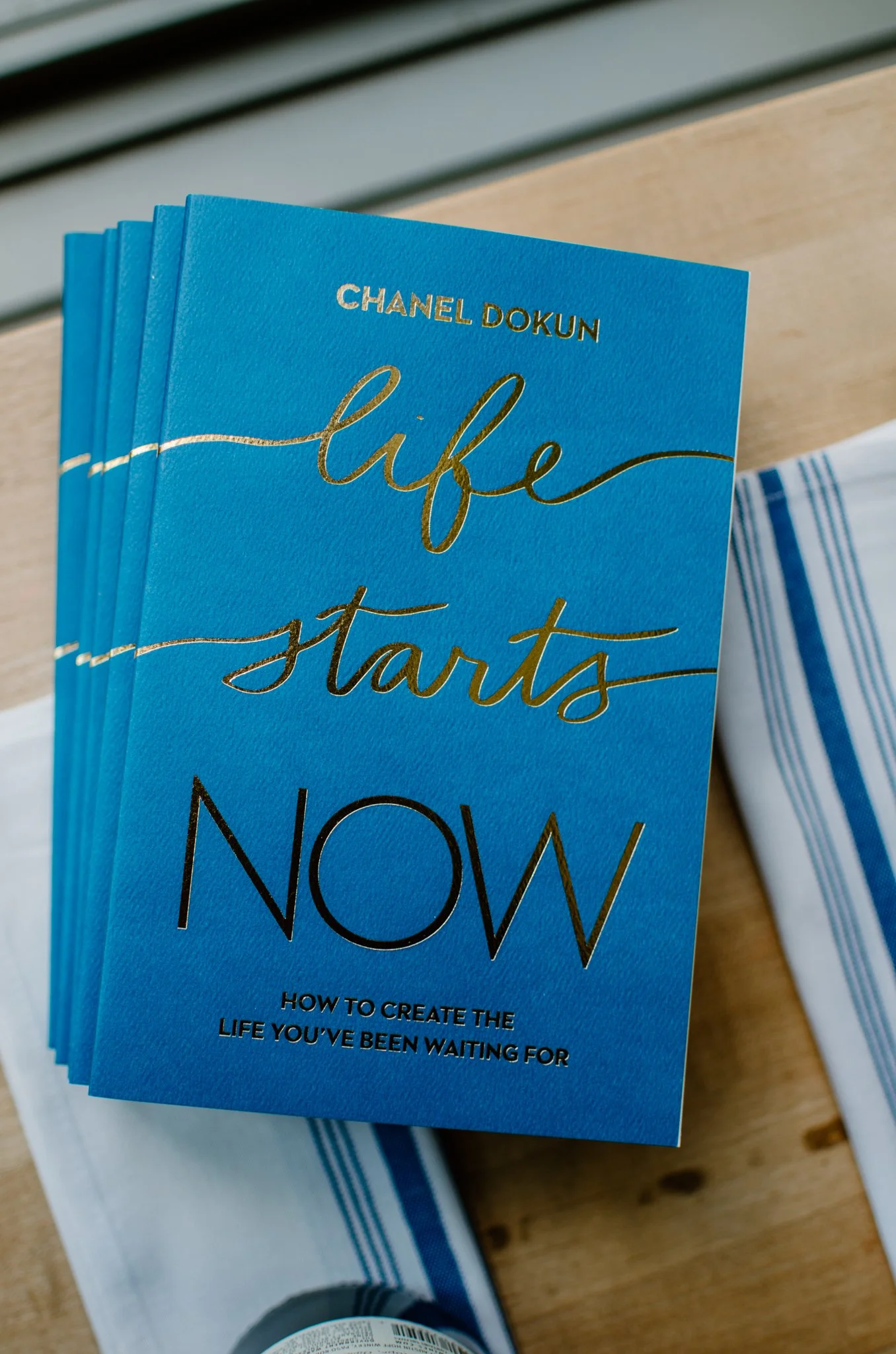 A blue book titled "Life Starts Now" by Chanel Dokun with subtitle "How to Create the Life You've Been Waiting For," displayed on a wooden surface, with another striped item partially visible in the background.