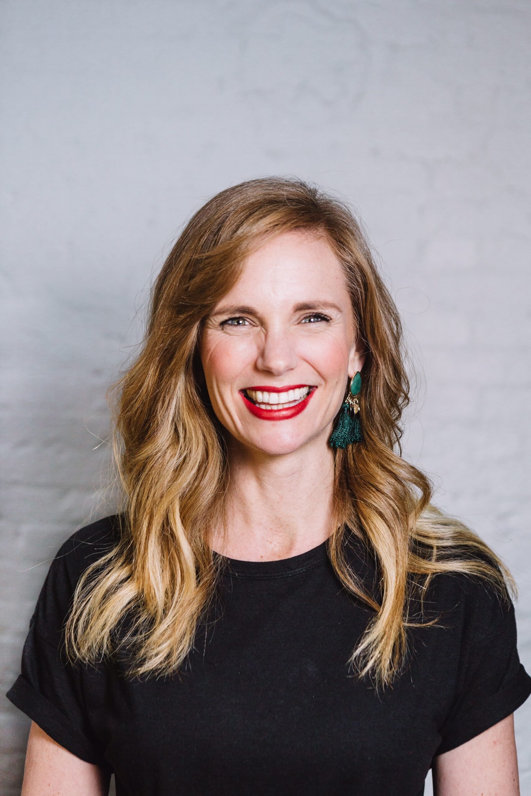 A smiling woman with long, wavy blonde hair, wearing red lipstick, teal earrings, and a black shirt photographed indoors against a plain light gray wall.