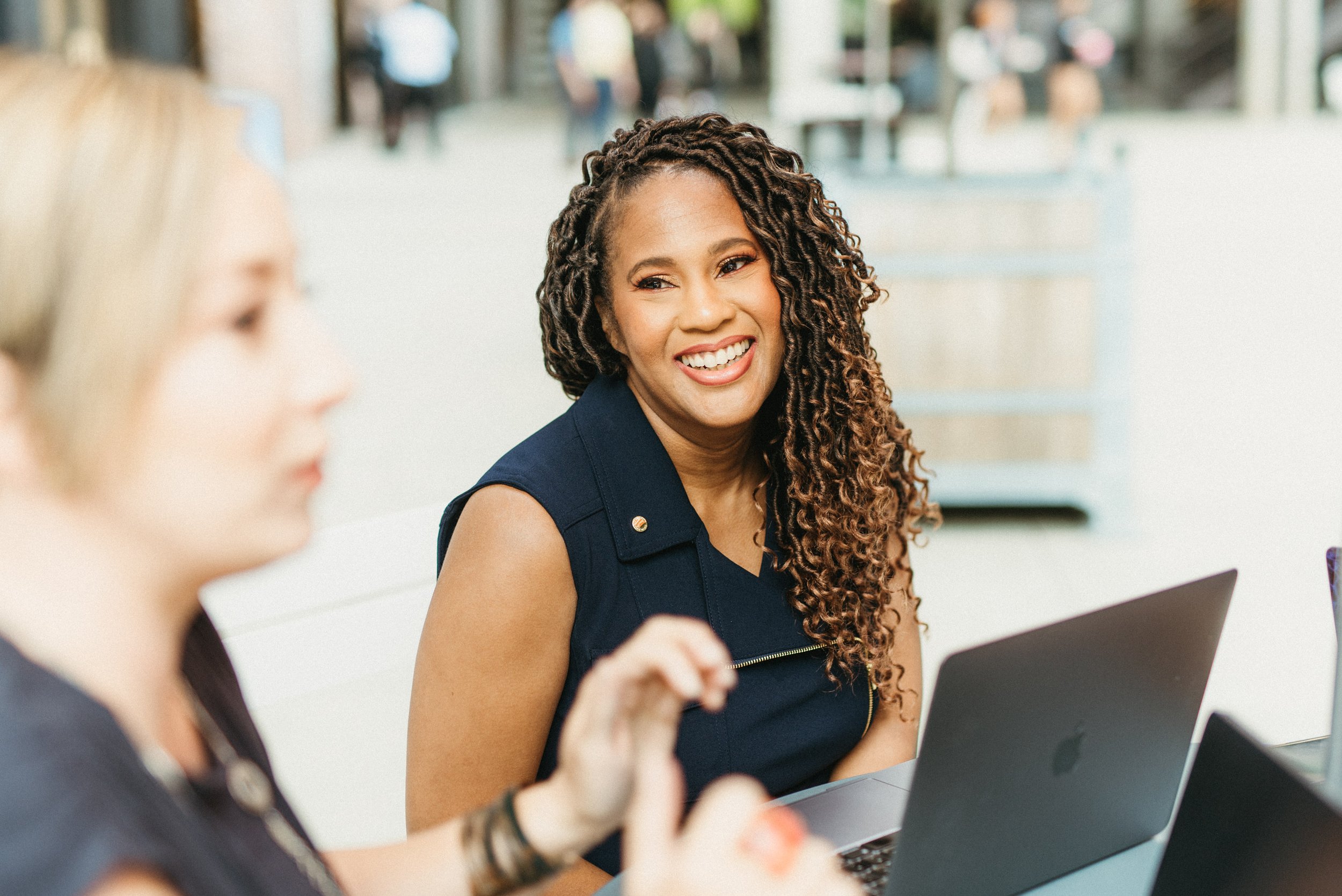 Smiling woman with curly hair sitting in a modern office, engaged in conversation with a blurred colleague in the foreground, both working on laptops.