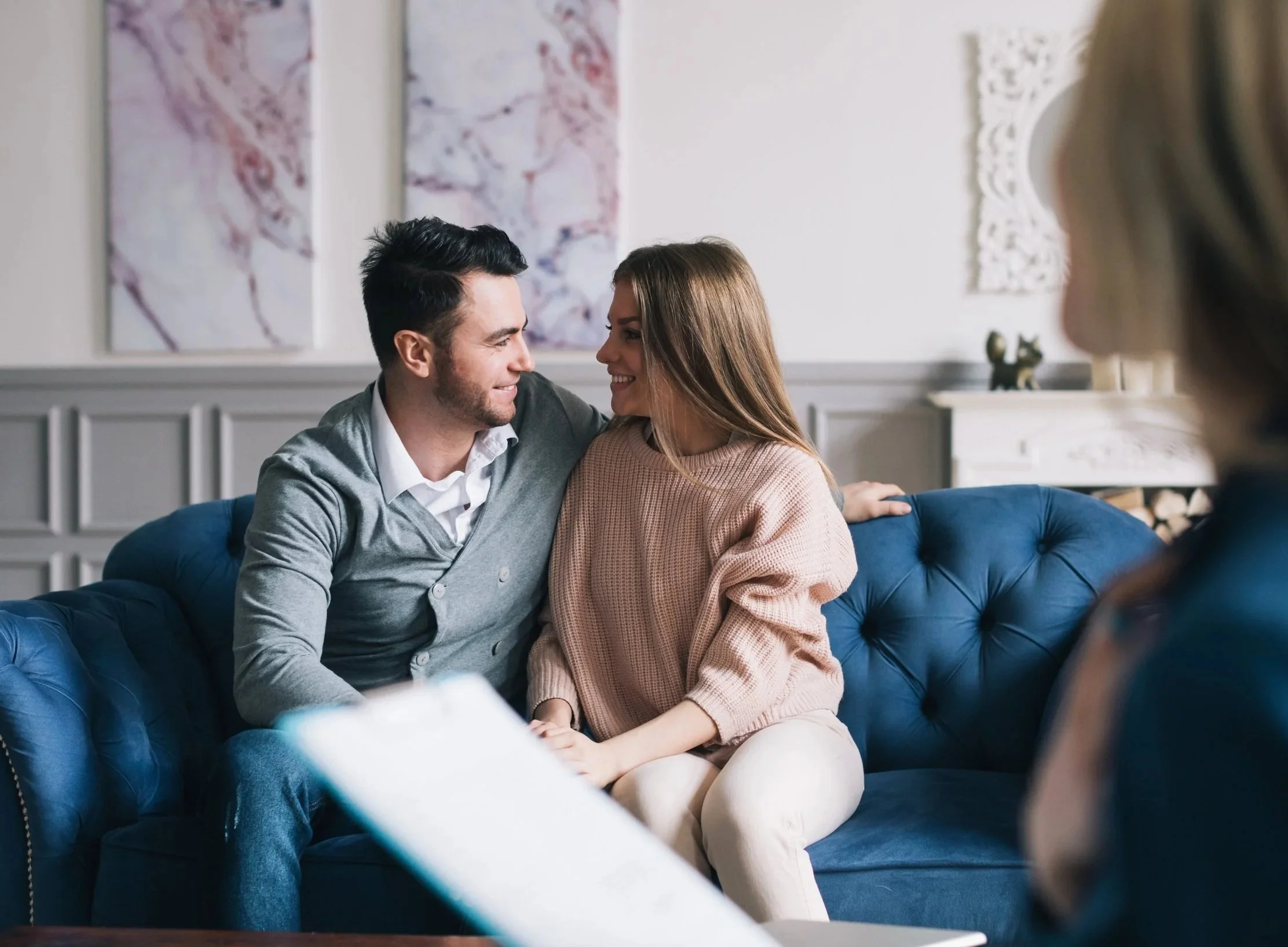 A couple is sitting closely together on a blue velvet couch, smiling and looking into each other's eyes, with a woman in a beige sweater and a man in a gray cardigan. They are in a living room decorated with artwork and a white wall with ornate decor.