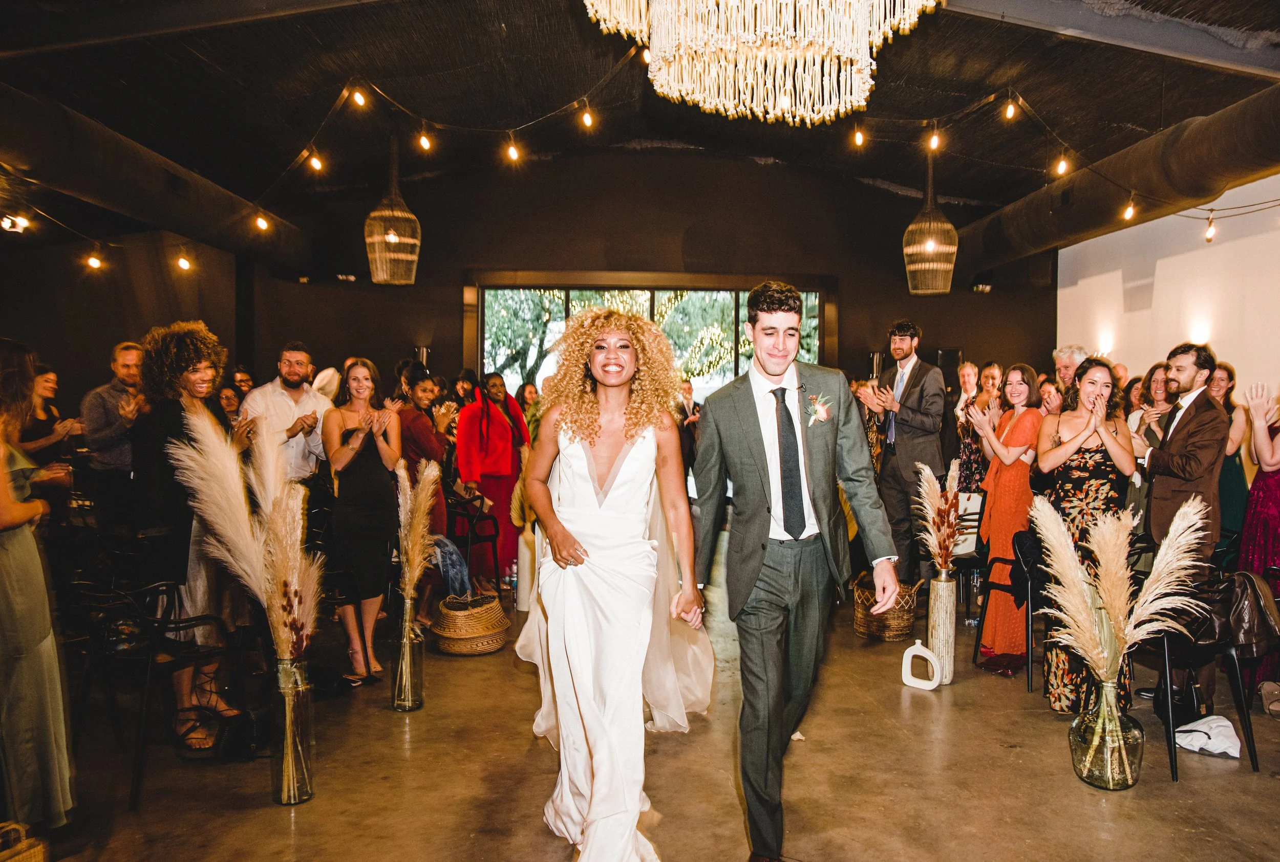 A bride and groom walk hand in hand down the aisle at their wedding reception as guests smile and applaud. The reception is decorated with string lights, warm lighting, and pampas grass centerpieces.